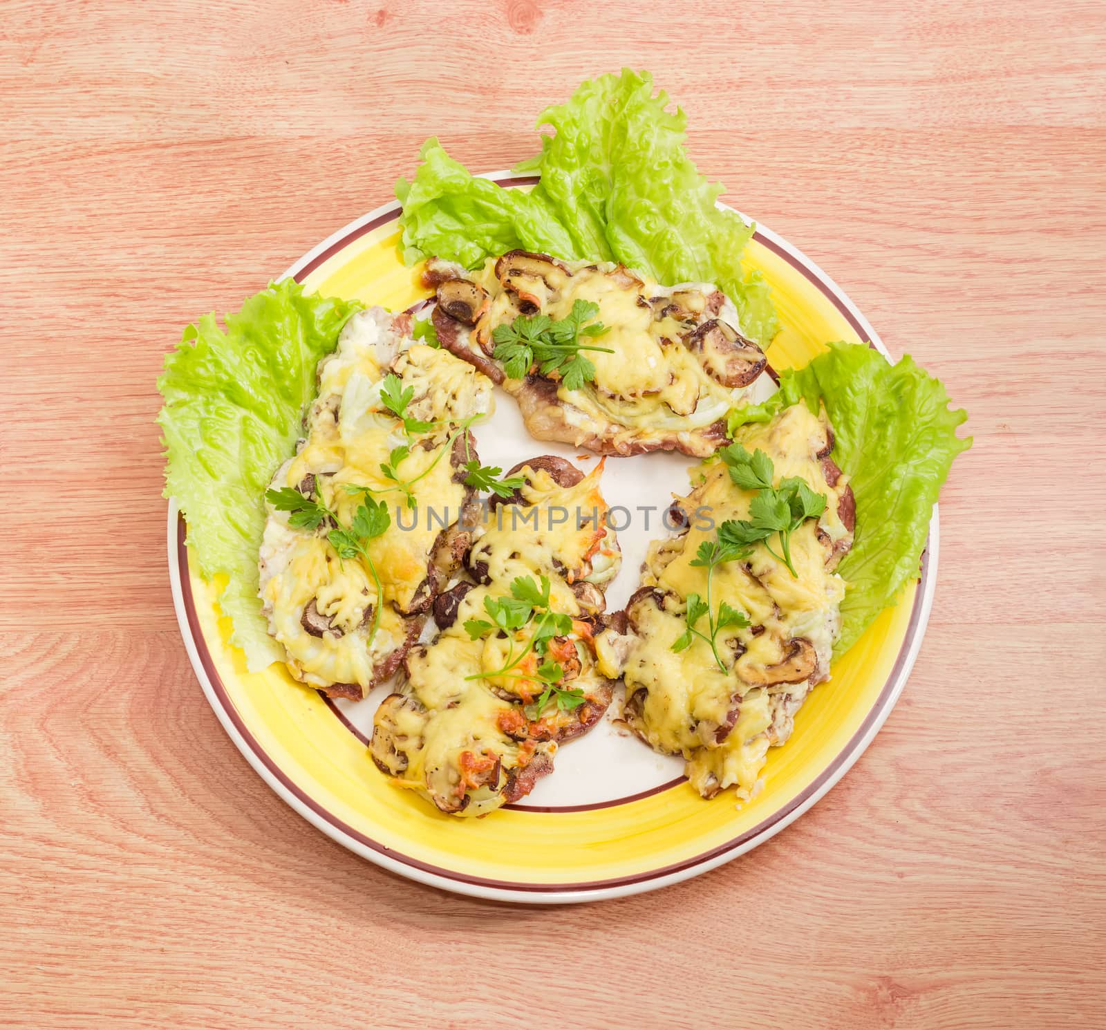 Top view of several pork chops baked with onion, mushrooms and cheese and decorated with twigs of the parsley and lettuce leaves on the yellow and white dish on a wooden surface
