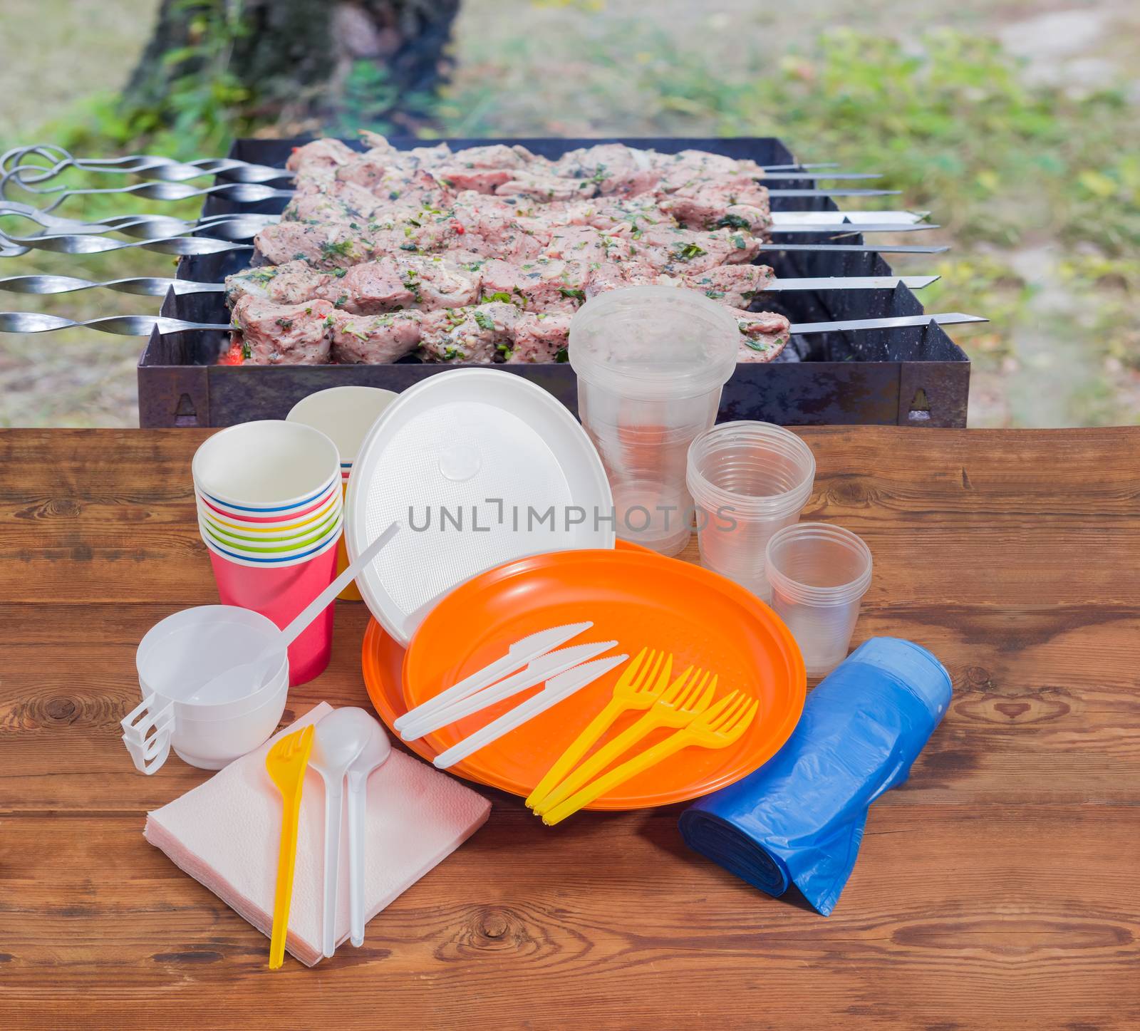 Different disposable plastic and paper cutlery, paper napkins and roll of disposable garbage bags on the old wooden planks against the background of charcoal grill with grilled skewered meat outdoors
