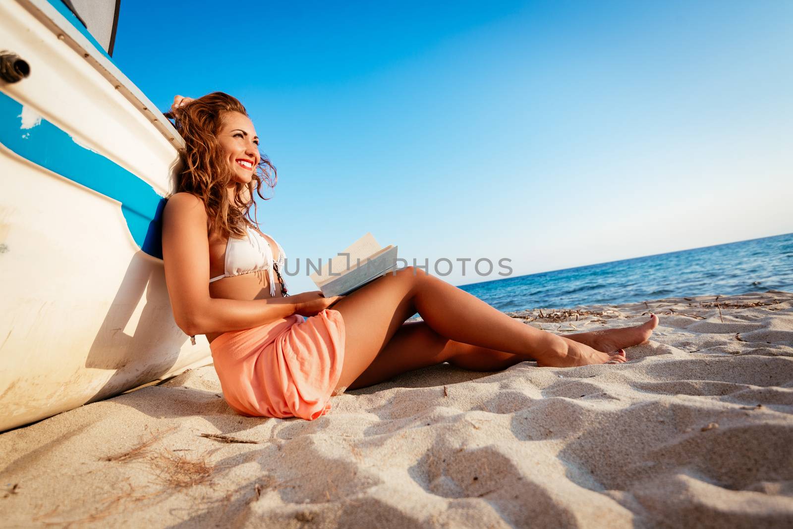 Young woman reading a book on the sandy beach.  She is sitting next to boat and looking away with smile on her thinking face.