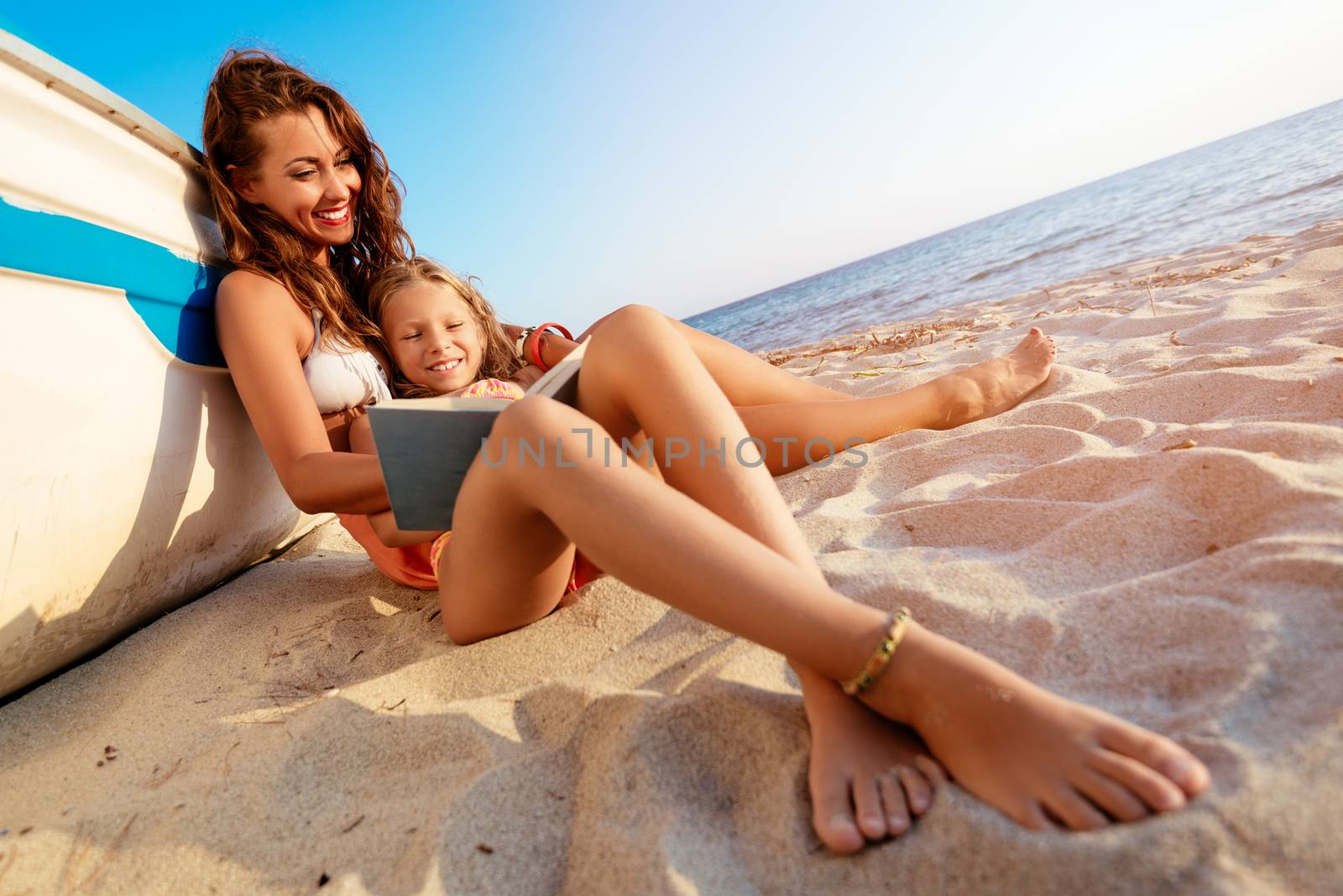 Beautiful little girl and young woman reading a book on the sandy beach. She is sitting next to boat with smile on her face.