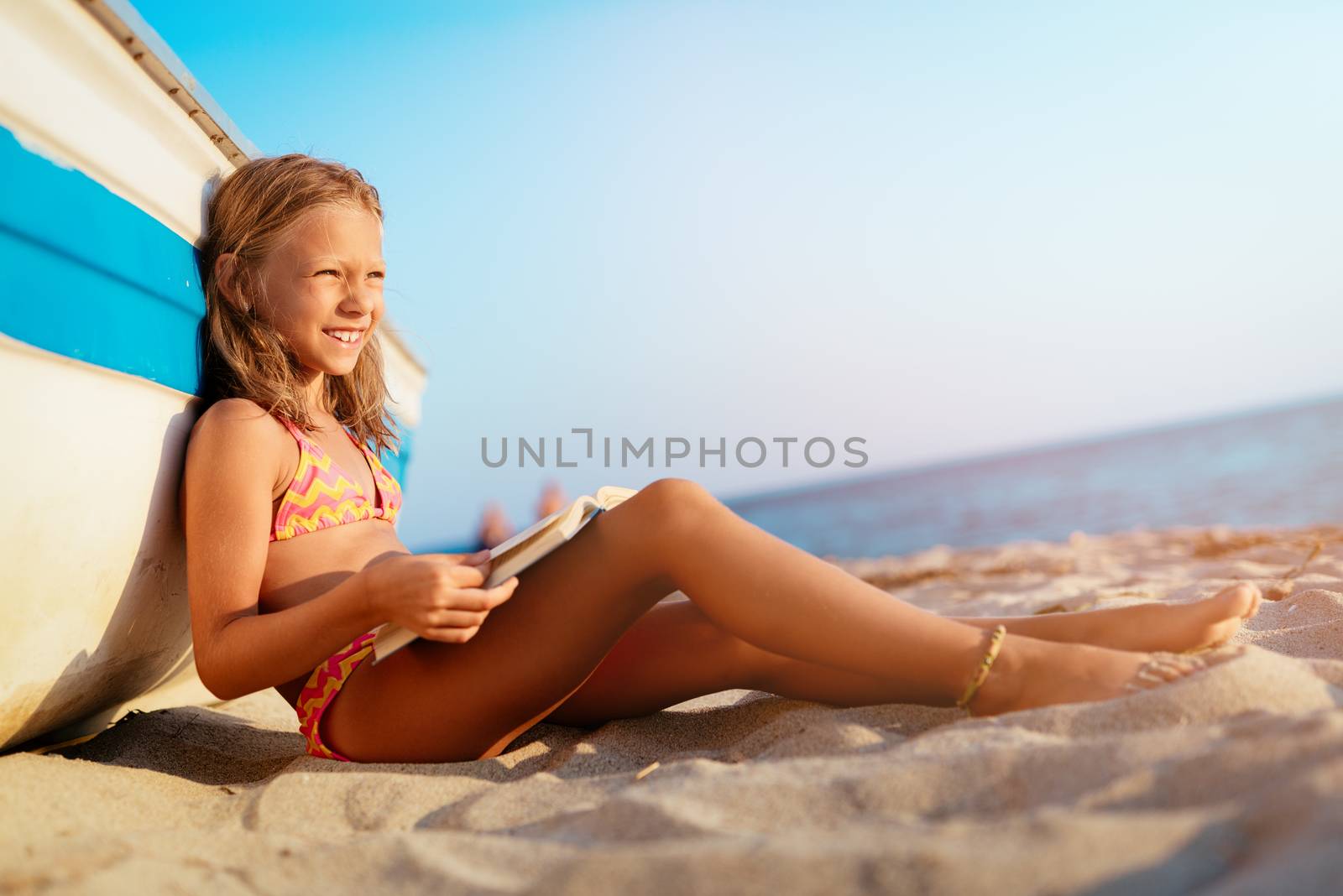 Beautiful little girl reading a book on the sandy beach.  She is sitting next to boat and looking away with smile on her thinking face.