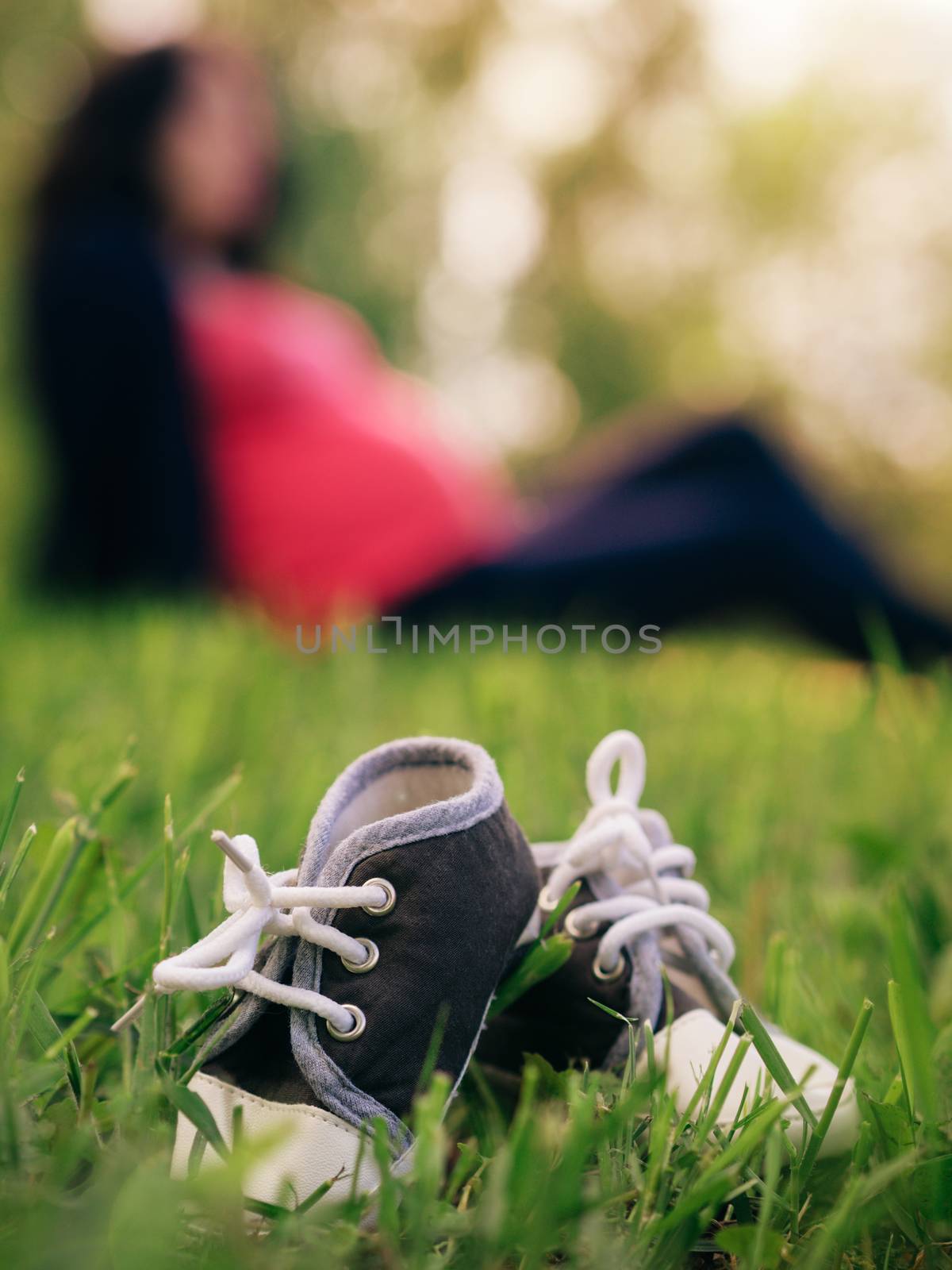 Close up view of little children shoes in grass and blurred silhouette of pregnant woman on background. Shallow DOF, focus on foreground. Pregnancy concept. Copy space. Vertical.