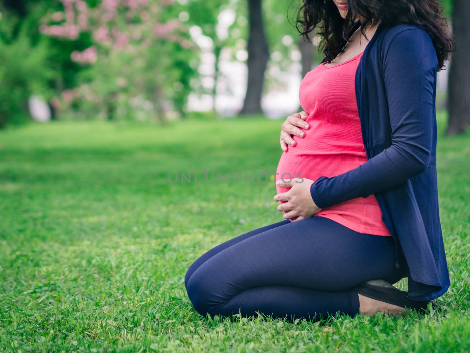 Close up view of pregnant woman siting on grass in park. Hand on abdomen. Copy space.