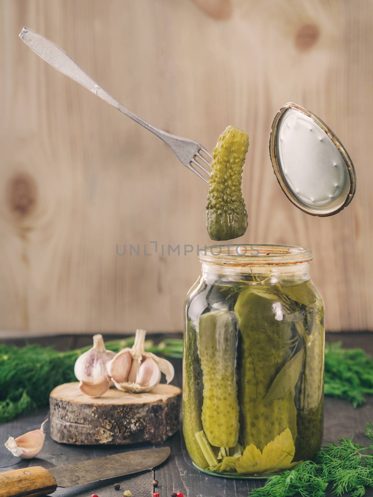 The fork holds a pickled cucumber that just has been taken from glass jar. Pickled cucumbers on wooden background. Pickles in glass jar with garlic, peppercorn and fresh dill. Flying food.