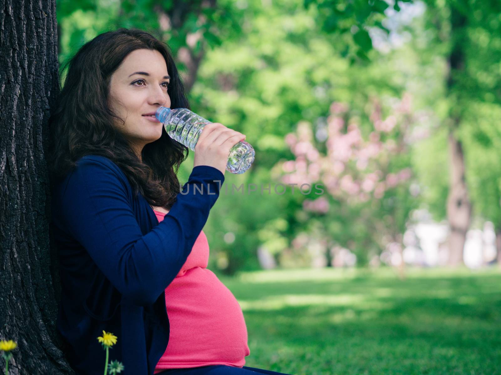 Pregnant woman drinks water from bottle outdoors. Copy space.
