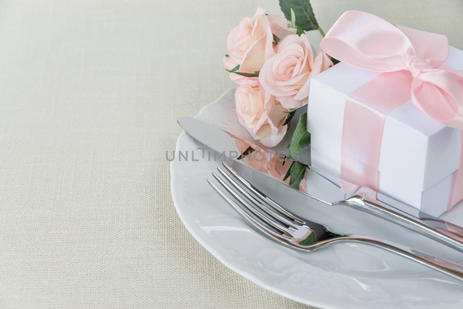 Beautiful decorated table with white plates, gift box with a pink ribbon, cutlery and pink rose flowers on tablecloths, with space for text