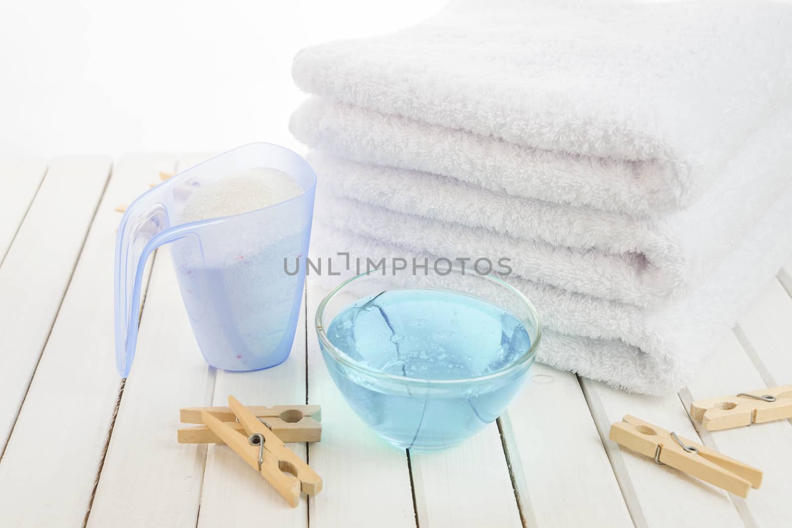 Stack of three white fluffy bath towels, washing powder in measuring cup, blue fabric softener in a transparent glass bowl and wooden clothespins on the background of white boards