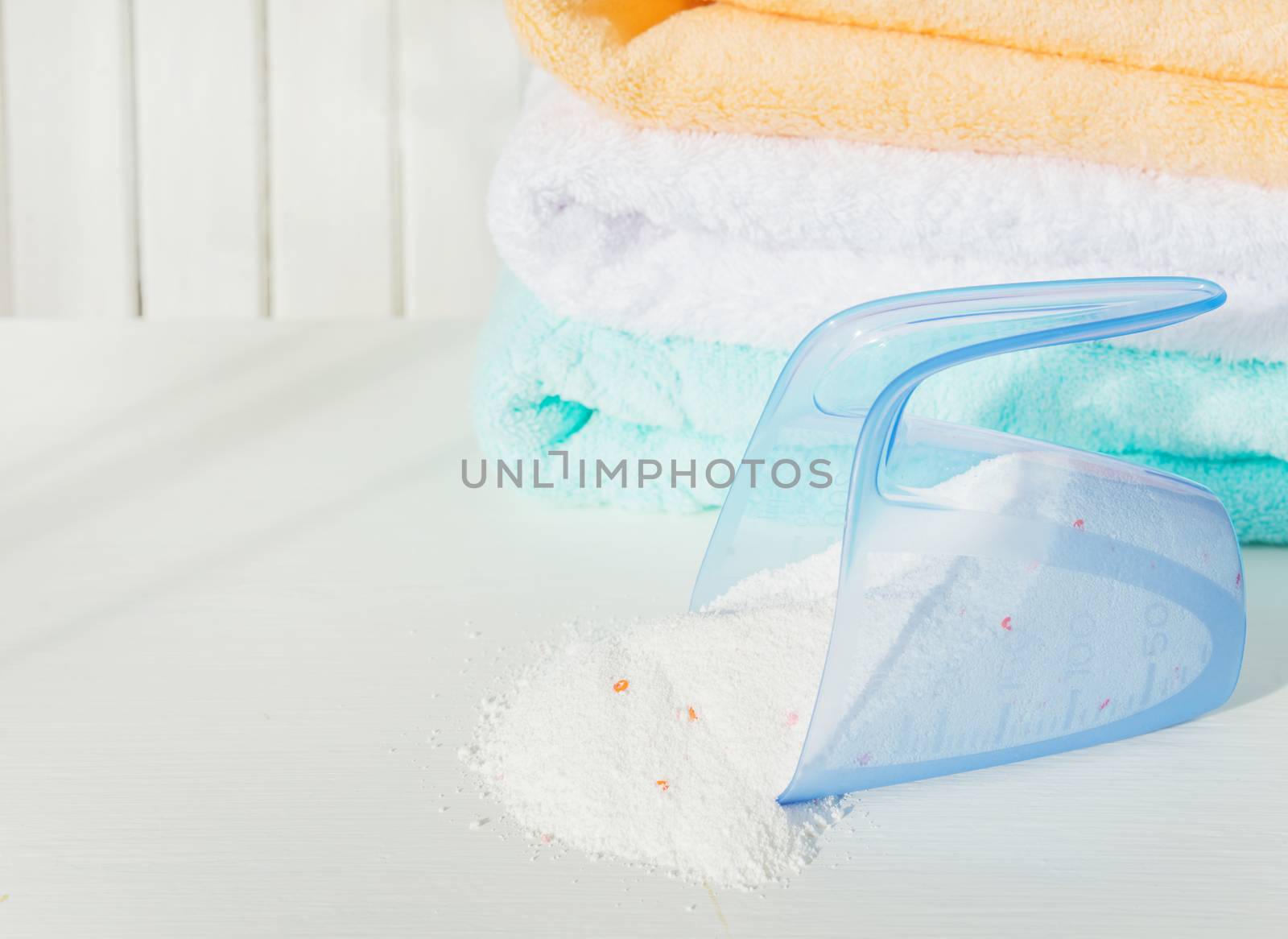 Stack of white, orange and blue fluffy bath towels and washing powder in measuring cup on the background of white boards