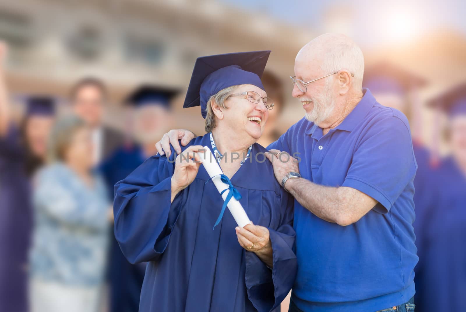 Senior Adult Woman In Cap and Gown Being Congratulated By Husband At Outdoor Graduation Ceremony. by Feverpitched