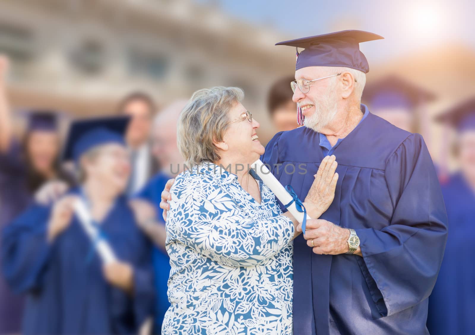 Senior Adult Male In Cap and Gown Being Congratulated By Wife At Outdoor Graduation Ceremony.