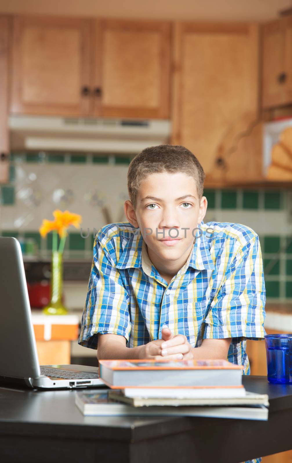 Calm attractive male teen leaning on kitchen counter with textbooks and laptop computer