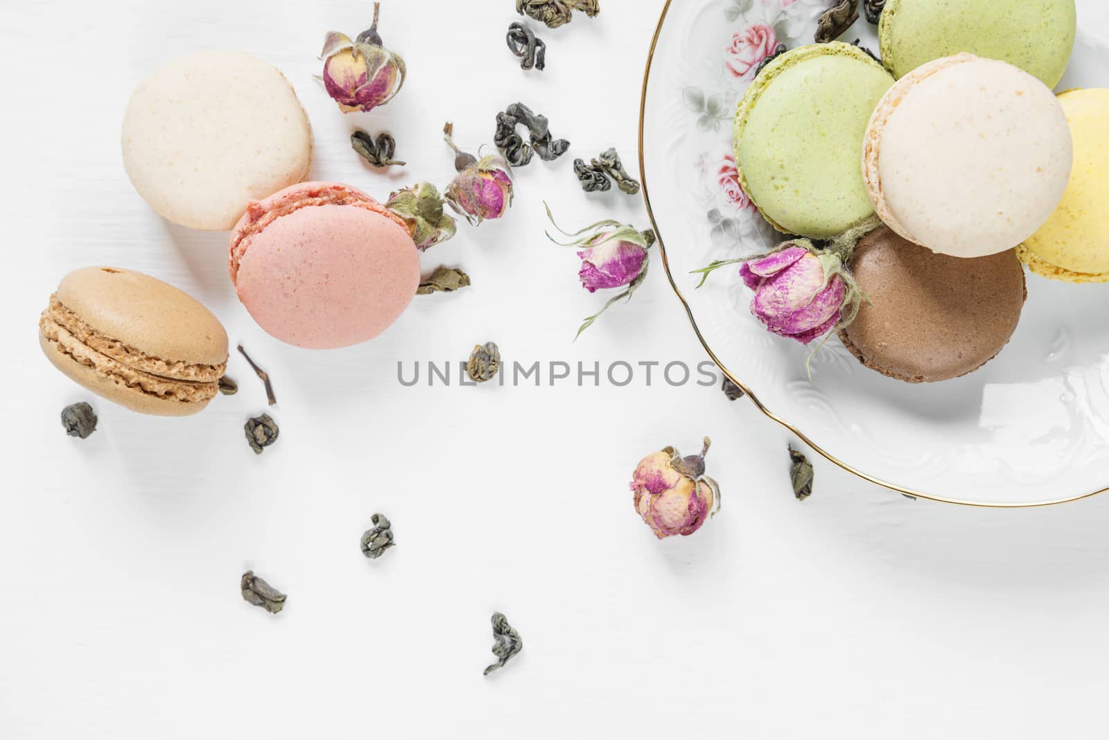 Multicoloured macaroons on beautiful porcelain plate, tea and dried rose buds on white background; top view, flat lay