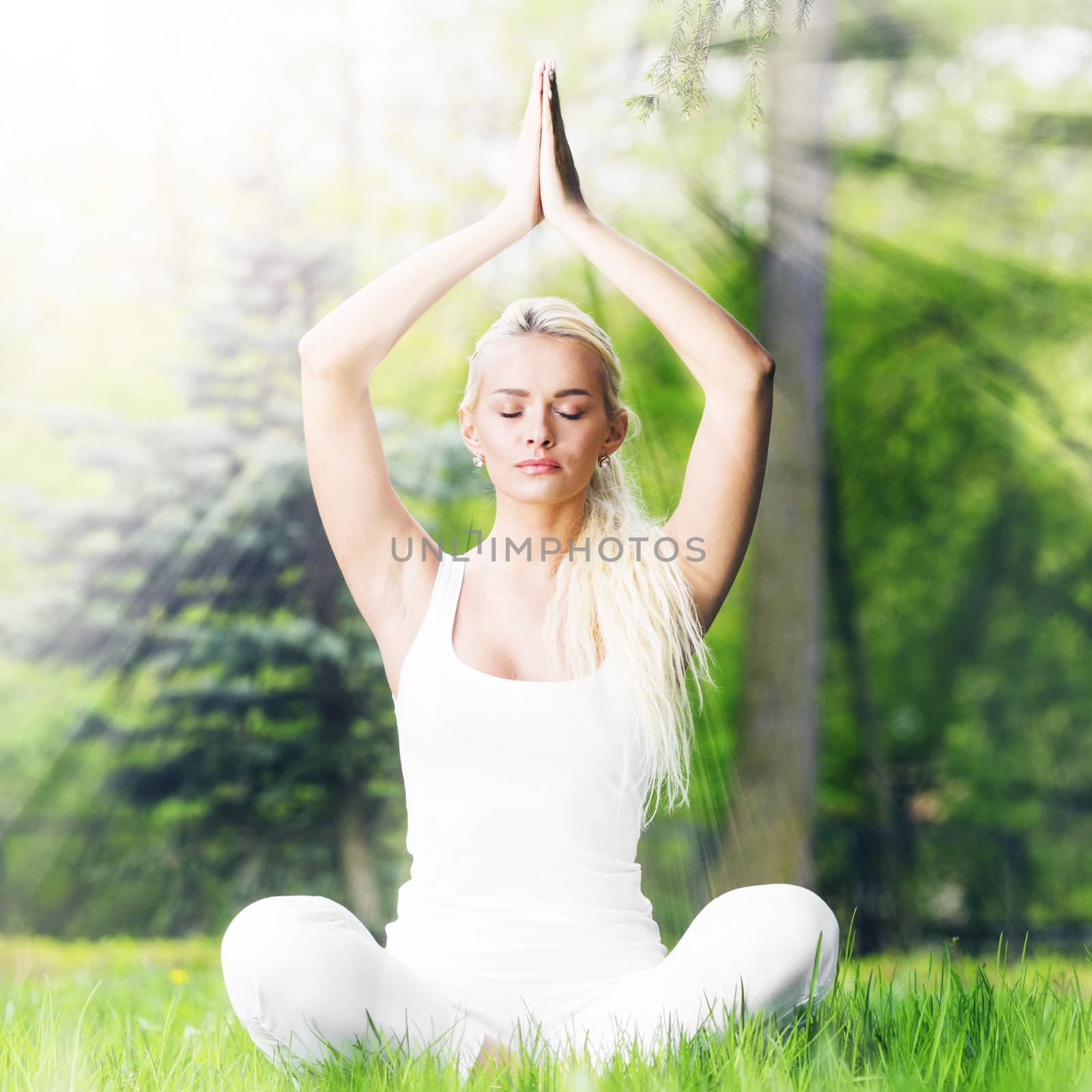 Beautiful young woman in white sportswear doing yoga lotus exercise in park in shiny rays of light
