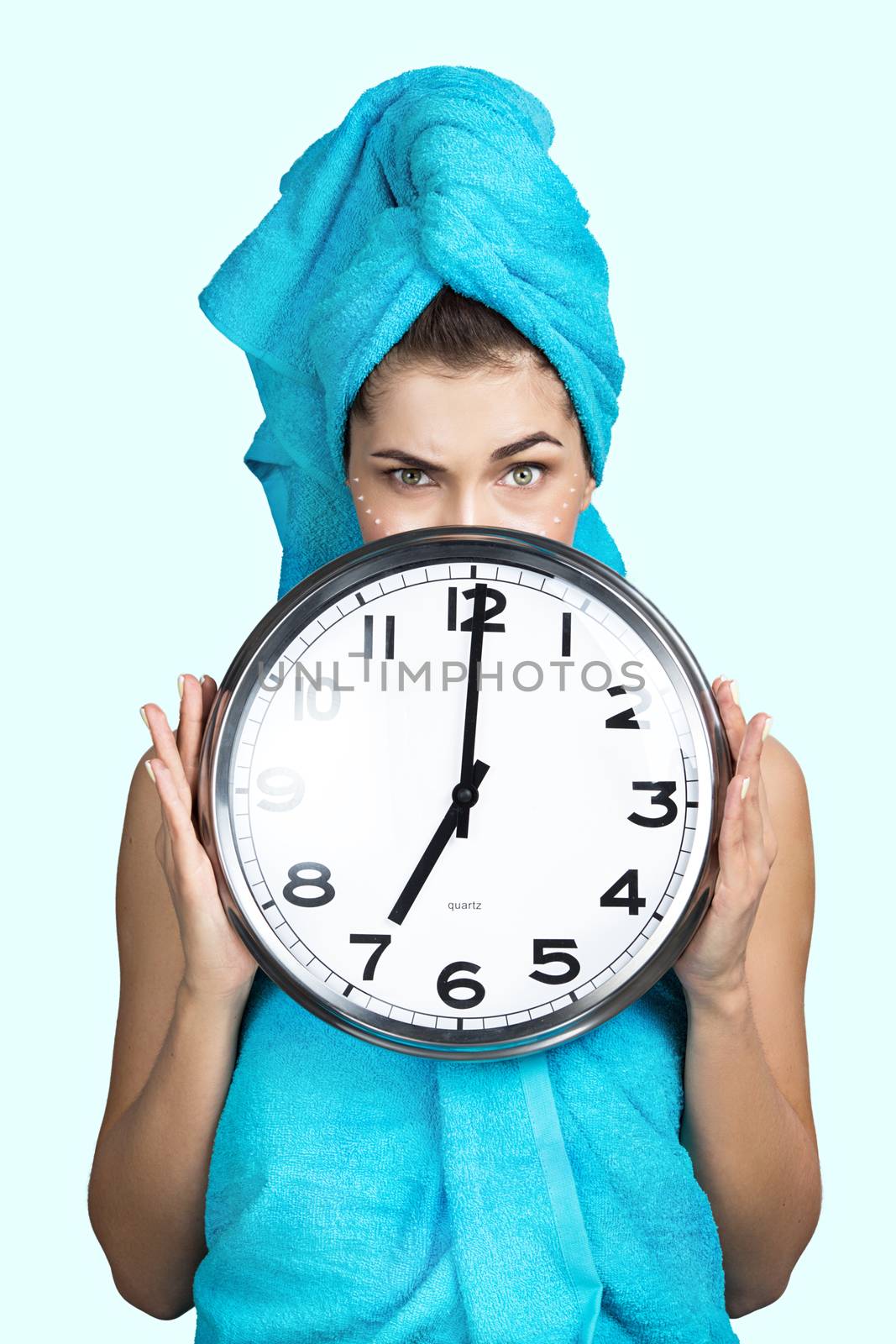 Woman in towel on head and creme on face holding wall clock showing 7, night or day creme skincare concept