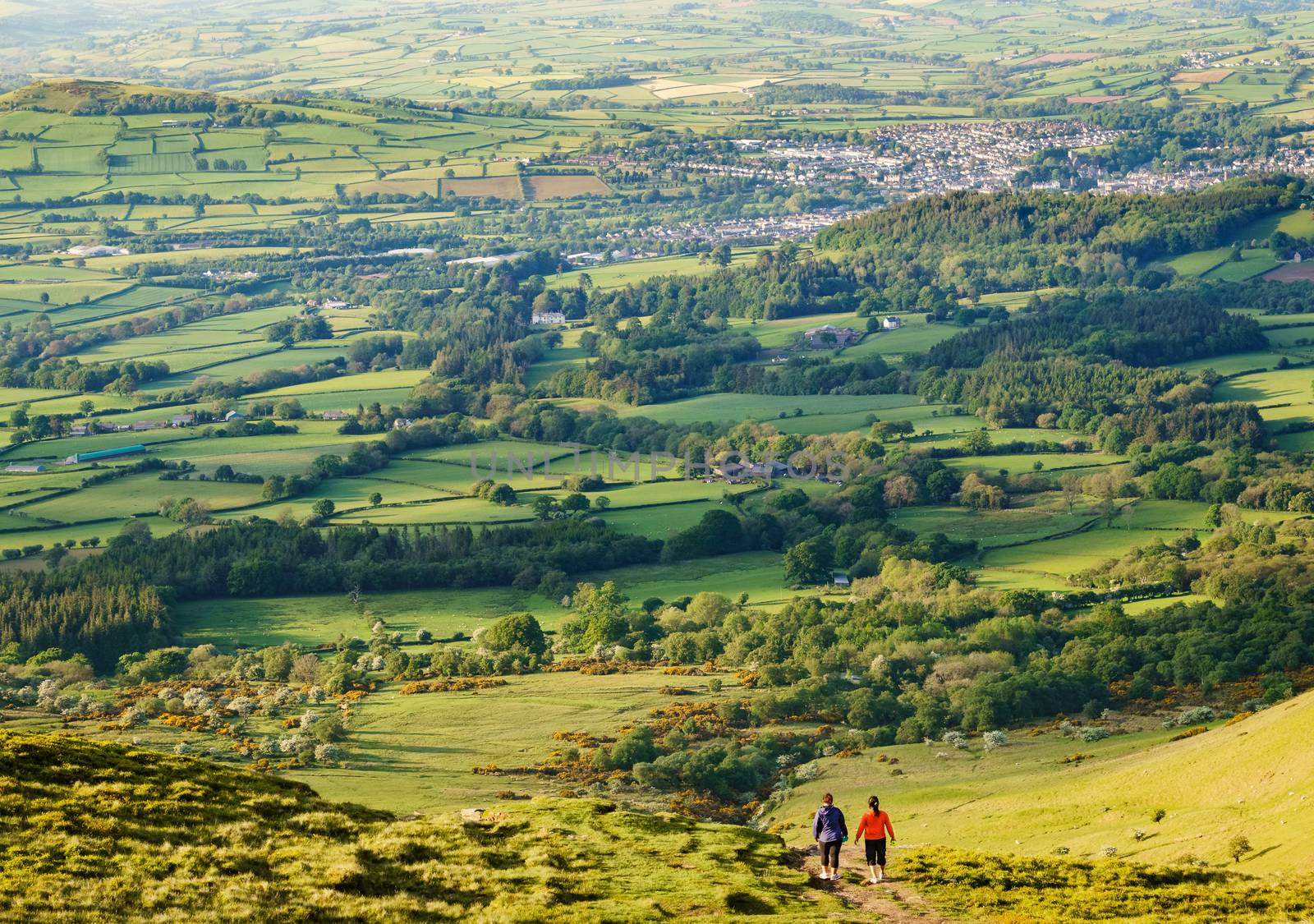 Two women walking away off of hill top in sports gear. Farmland and town in the distance. Taken in Wales during golden hour.