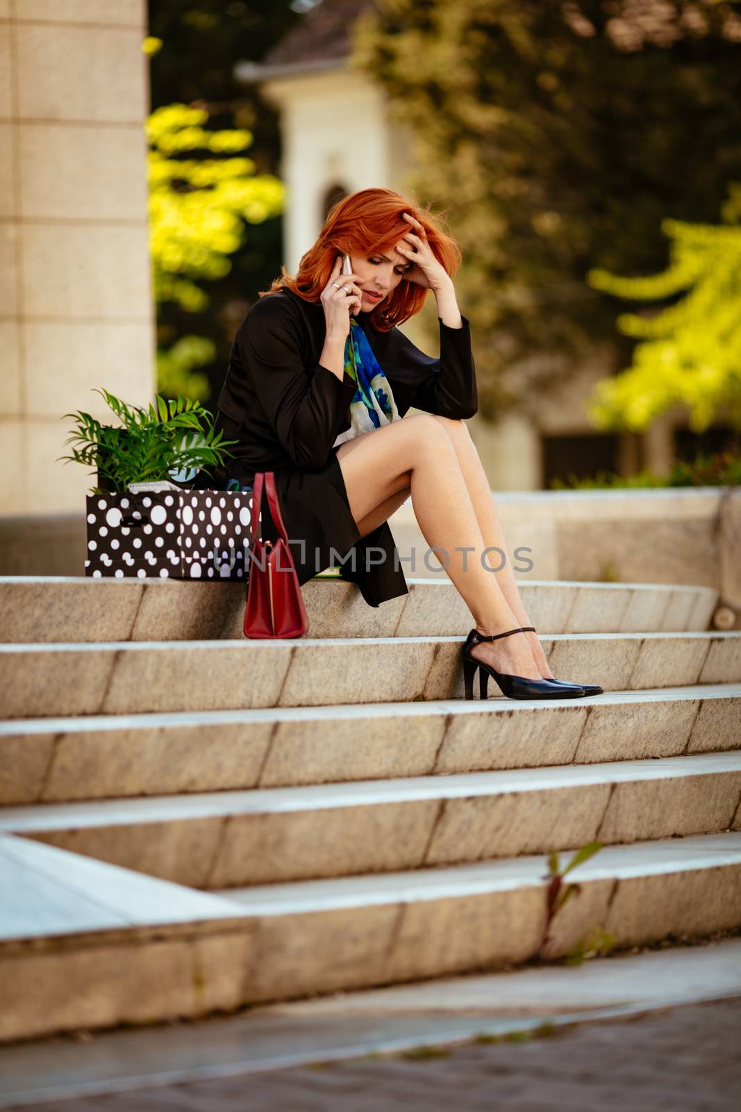 Serious pensive businesswoman sitting on stairs and talking on smartphone next to a box full of her personal belongings from the office just after she got fired. 