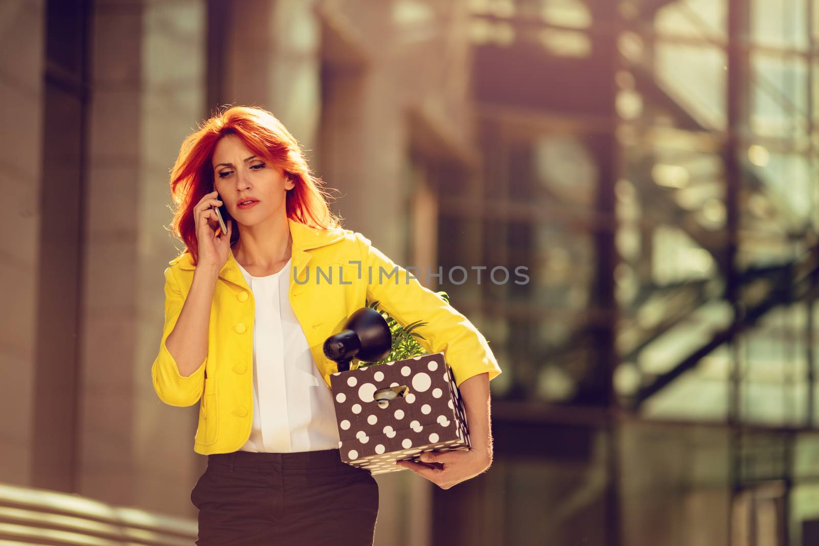 Serious pensive businesswoman walking in office district and talking on smartphone with a box full of her personal belongings from the office just after she got fired. 