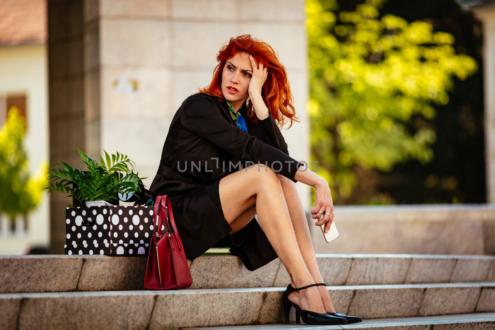 Serious pensive businesswoman sitting on stairs next to a box full of her personal belongings from the office just after she got fired. 