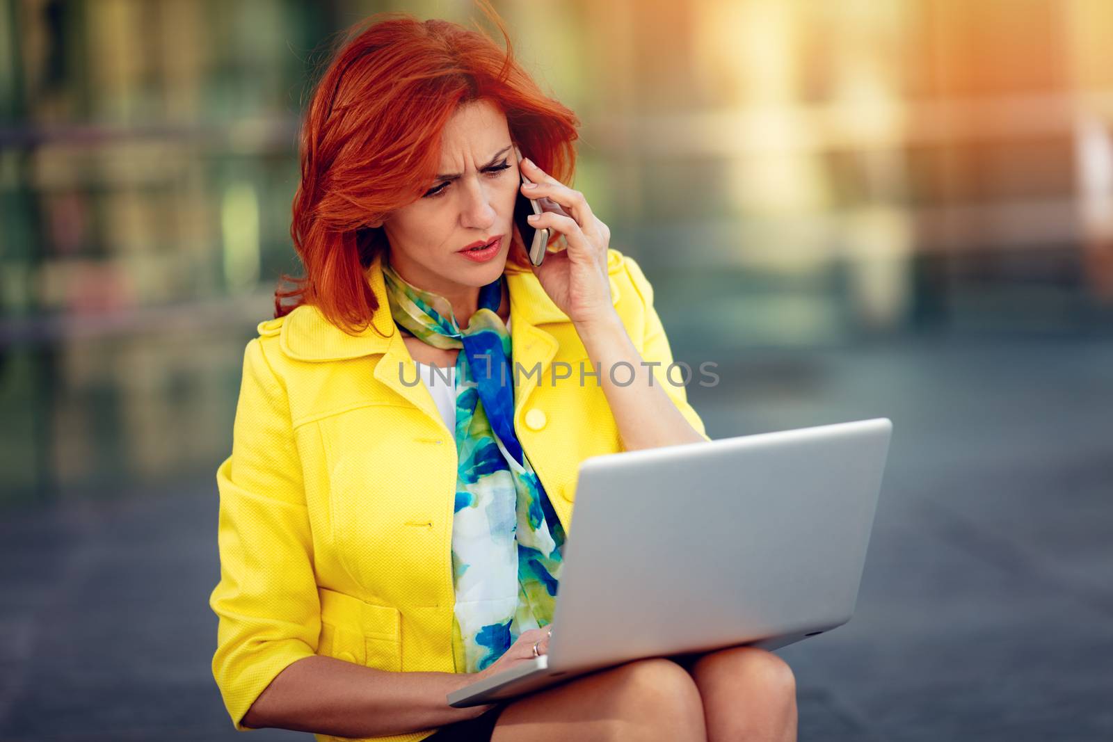 Serious businesswoman sitting on stairs in office district, working at the laptop and talking on smartphone.