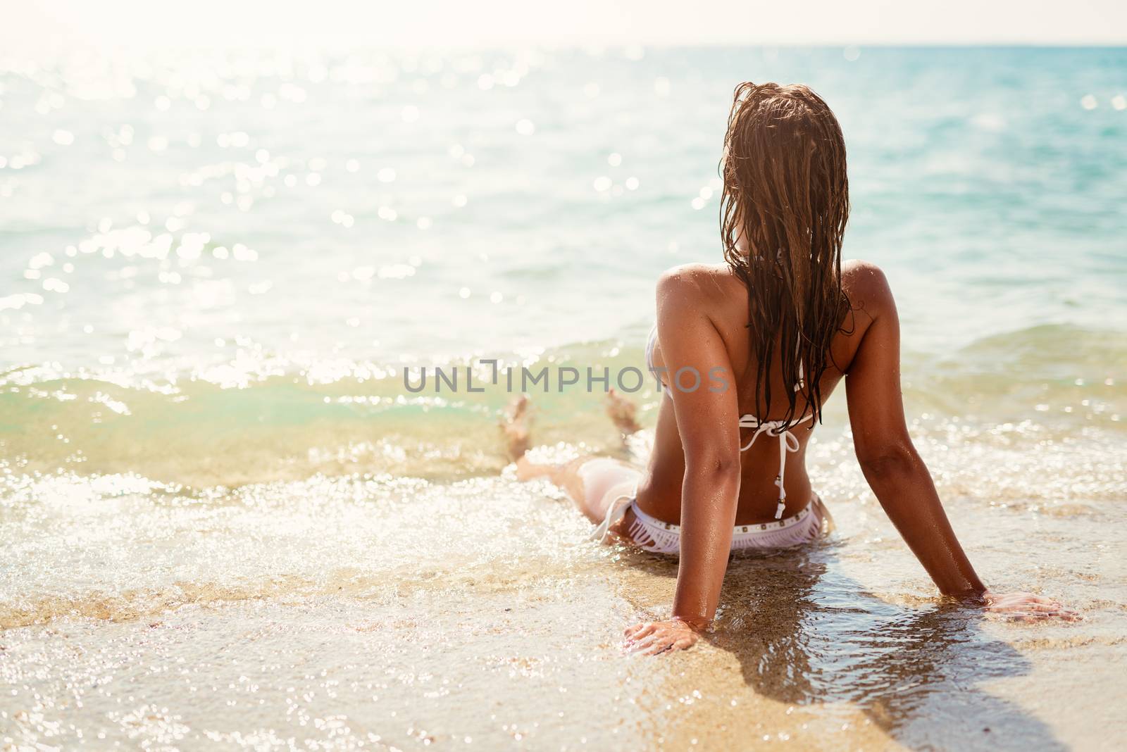 Rear view of a beautiful young woman enjoying on the beach. She is lounging and sunbathing on the sandy coast in the sea. 