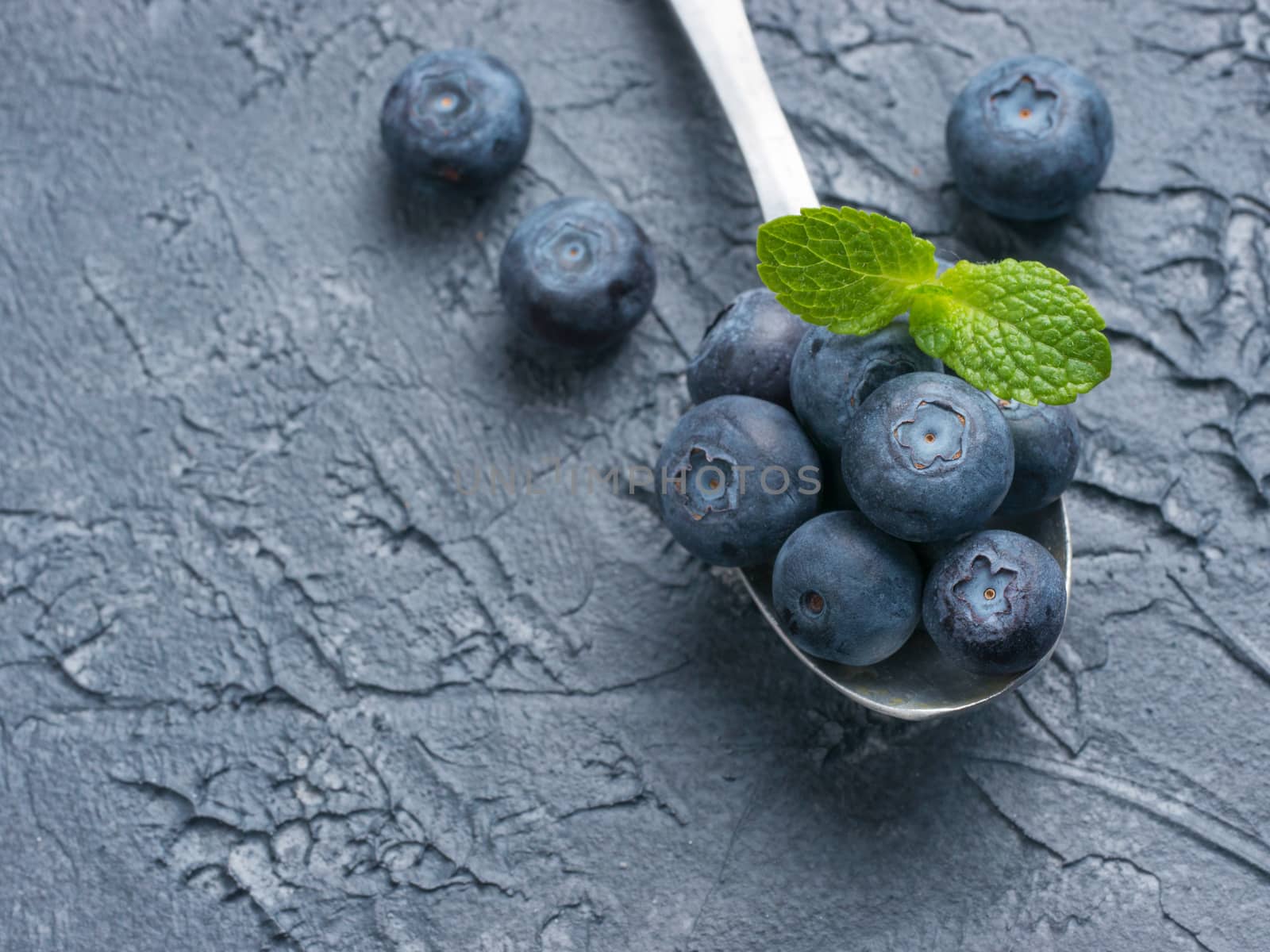 Freshly picked blueberries in spoon closeup. Ripe and juicy fresh blueberry with green mint leaves on textured concrete background. Bilberry on dark background with copyspace. Top view or flat lay