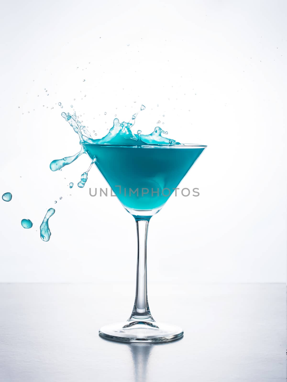 Blue cocktail in martini glass with ice cube splashing into liquid against white background. Blue curacao cocktail with splash on white.