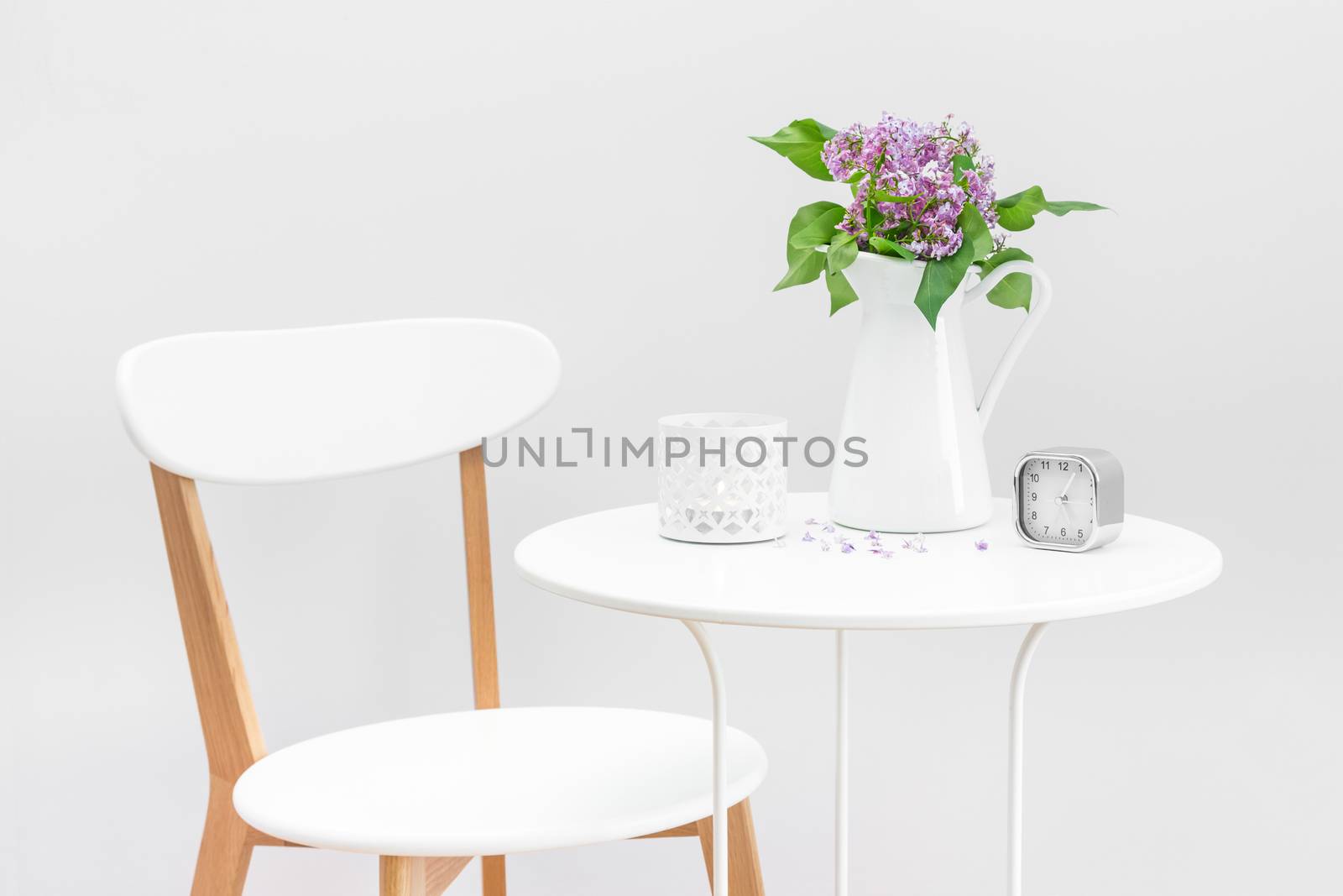 Elegant interior with white table and chair, decorated with a bouquet of purple lilacs, candle and clock.