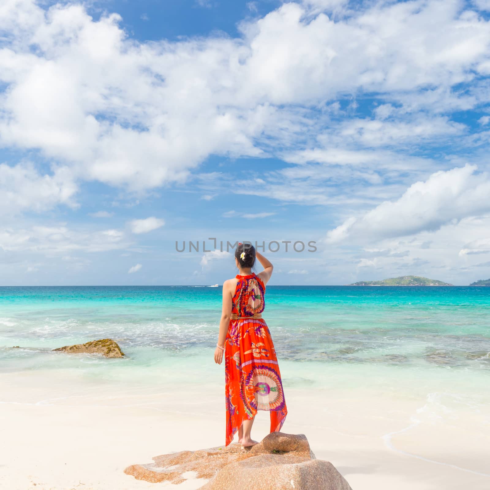 Traditinaly dressed local woman wearing long floral summer dress and hat looking at sea on Anse Patates beach, La Digue Island, Seychelles. Summer vacations on picture perfect tropical island concept.
