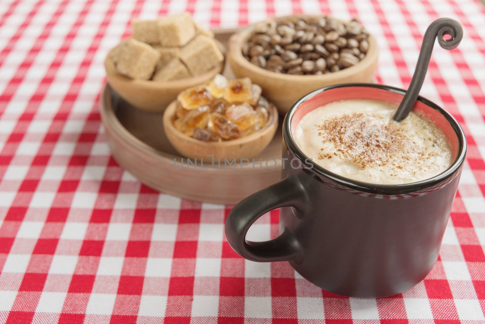 Black coffee with rich crema in a black ceramic mug, cane sugar, caramel and coffee beans in the wooden bowls are on a background of red and white checkered cloth