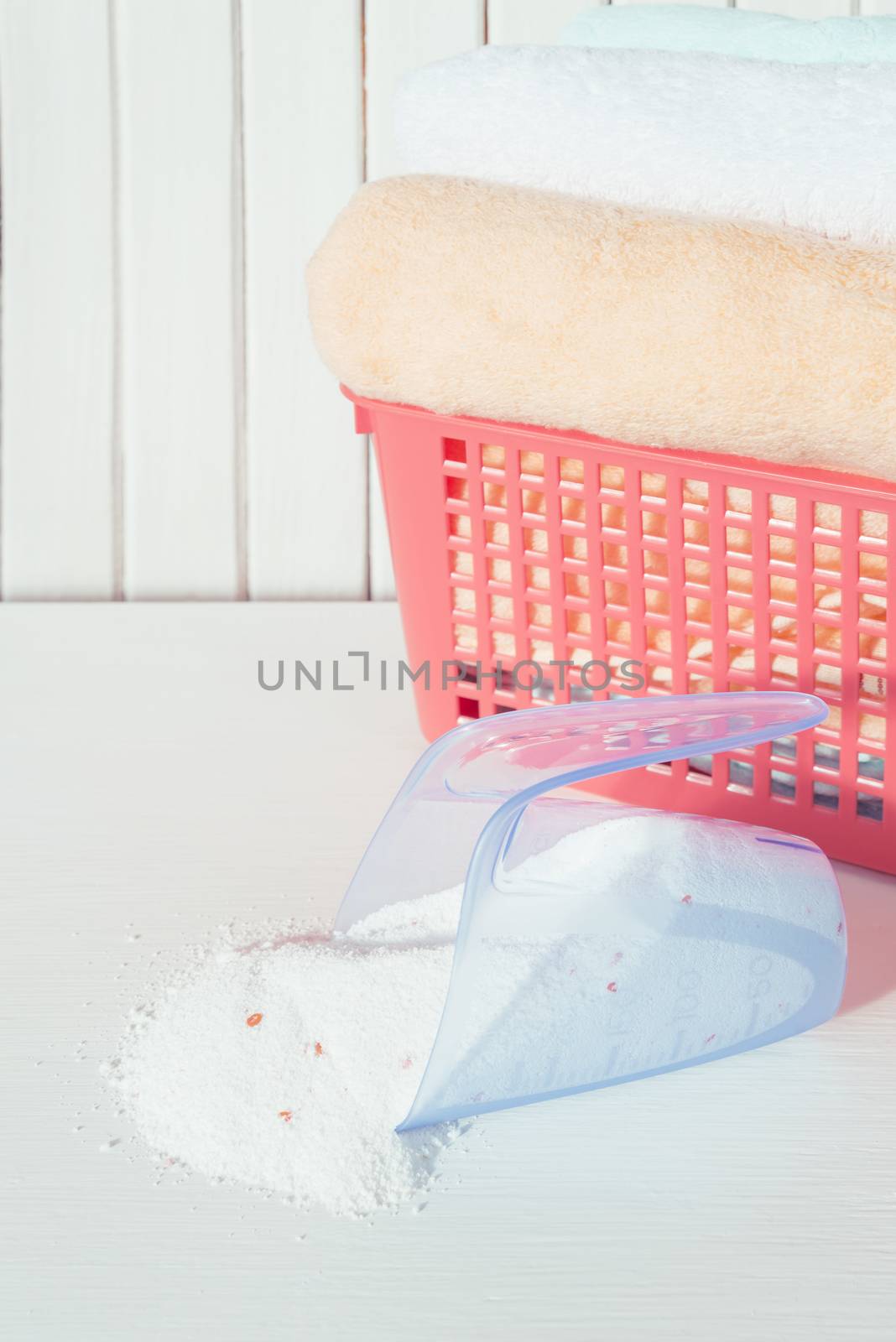 White, orange and blue fluffy bath towels in the red laundry basket and spilled washing powder in measuring cup on the background of white boards