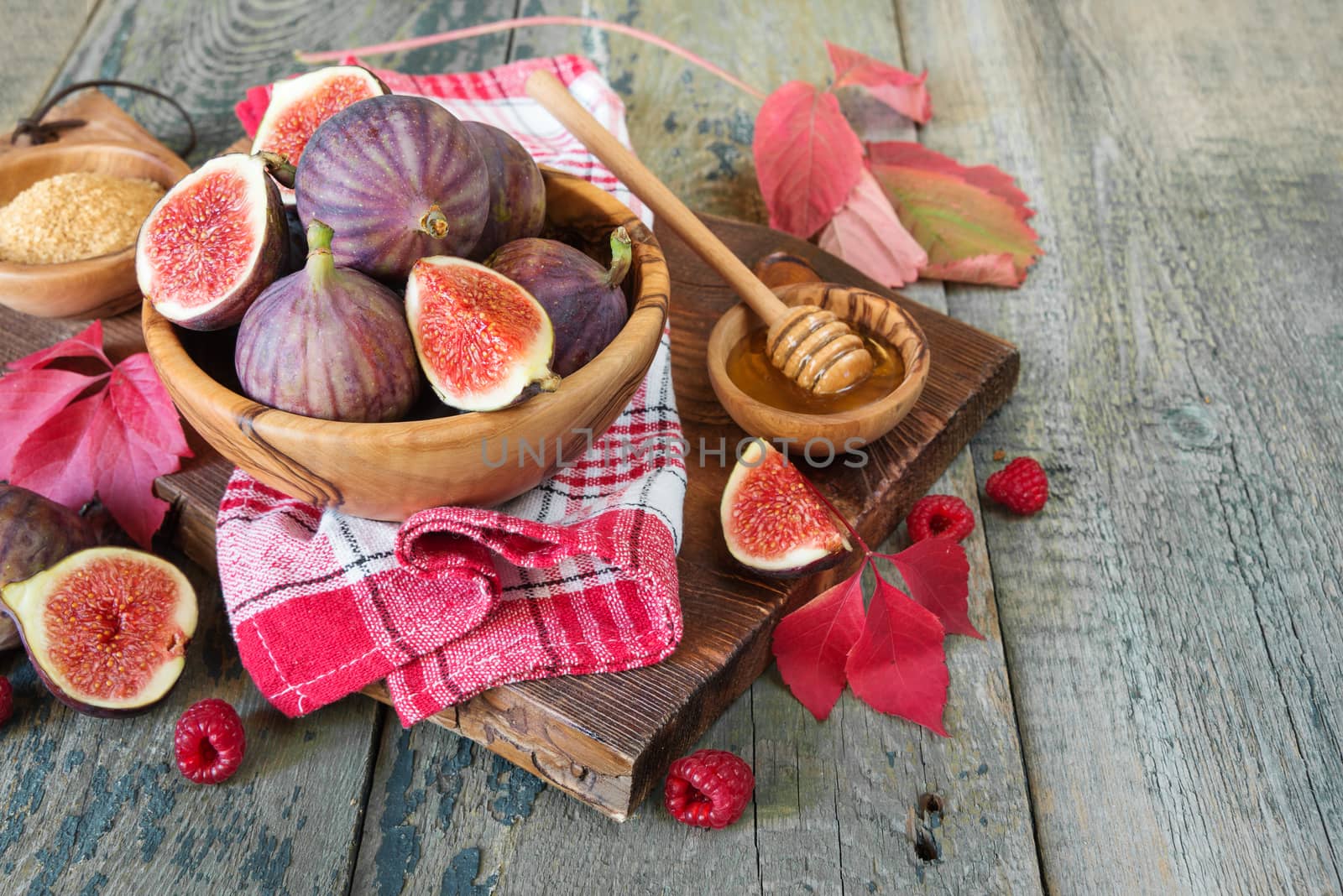 Ripe figs in a wooden bowl, red raspberry, cane sugar, honey and a checkered napkin on old cutting board as well as autumn leaves lie on the old wooden table