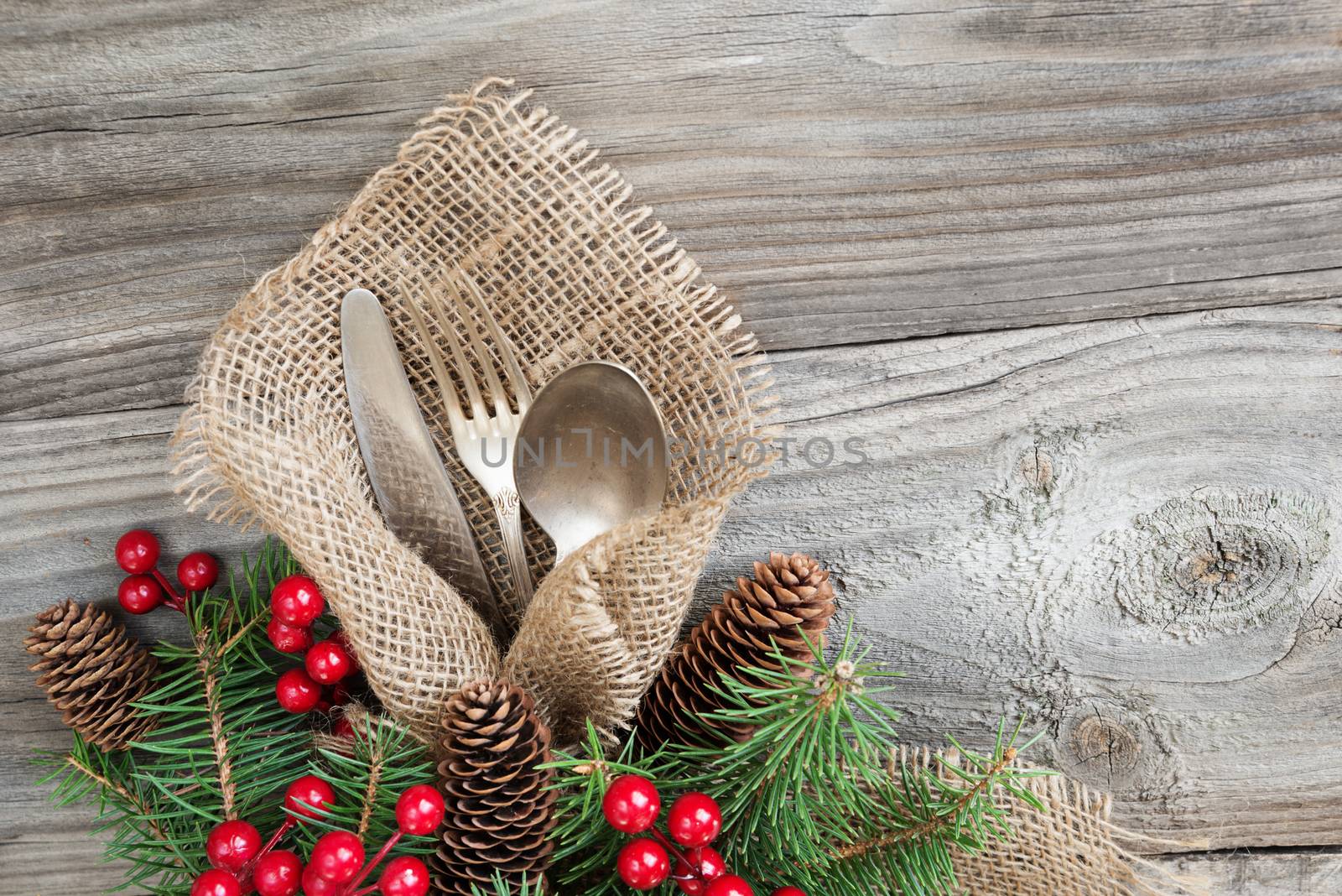 Christmas table with Christmas decoration: old knife, spoon and fork lie on the sacking napkin, as well as red holly berries and green spruce branch, which is located on an old wooden table, with space for text