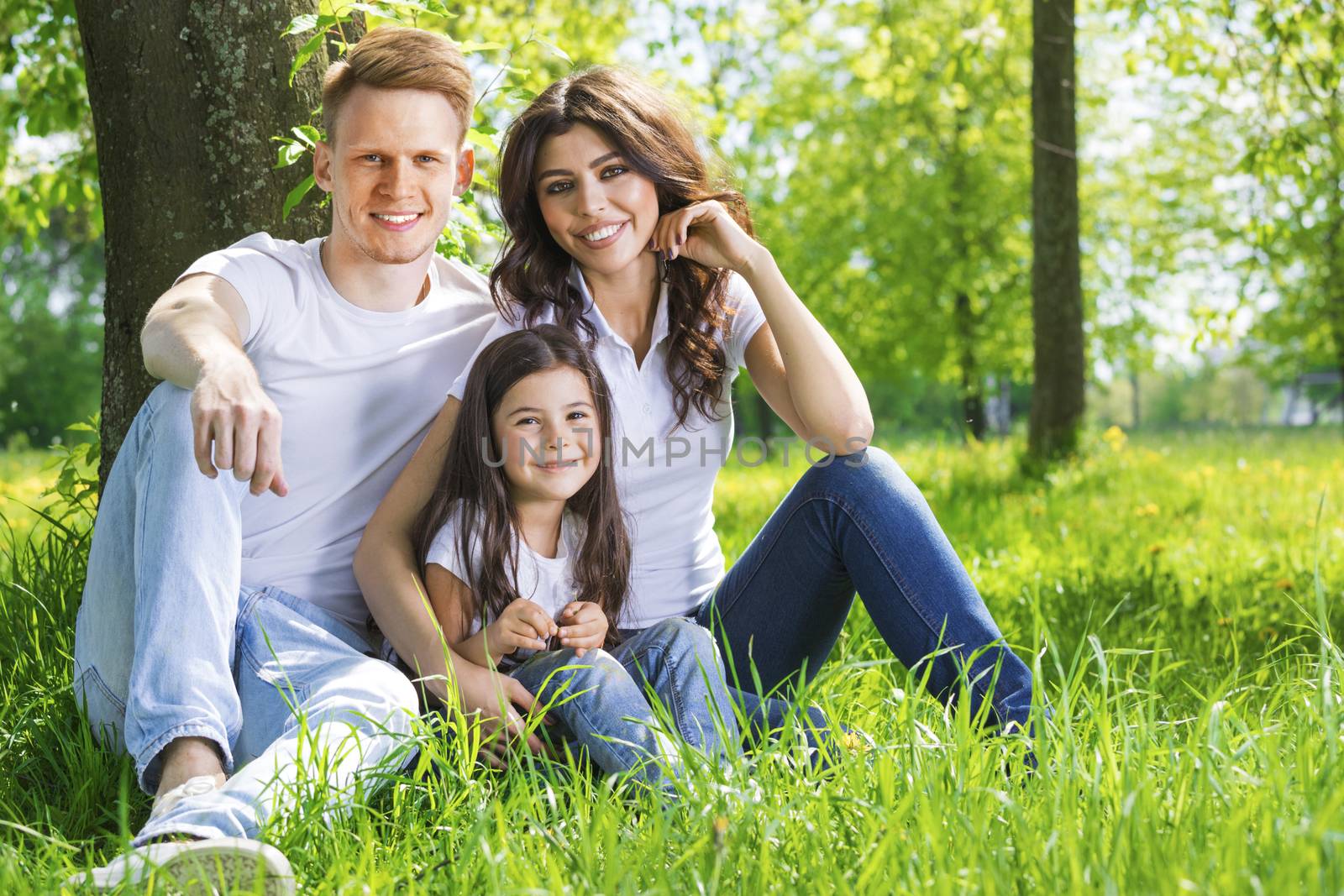 Portrait of happy family sitting on grass under a tree in park on a sunny day