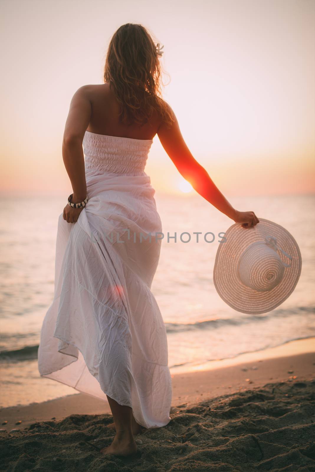 Rear view of a woman in white dress enjoying sunset on the beach and holding white hat. 
