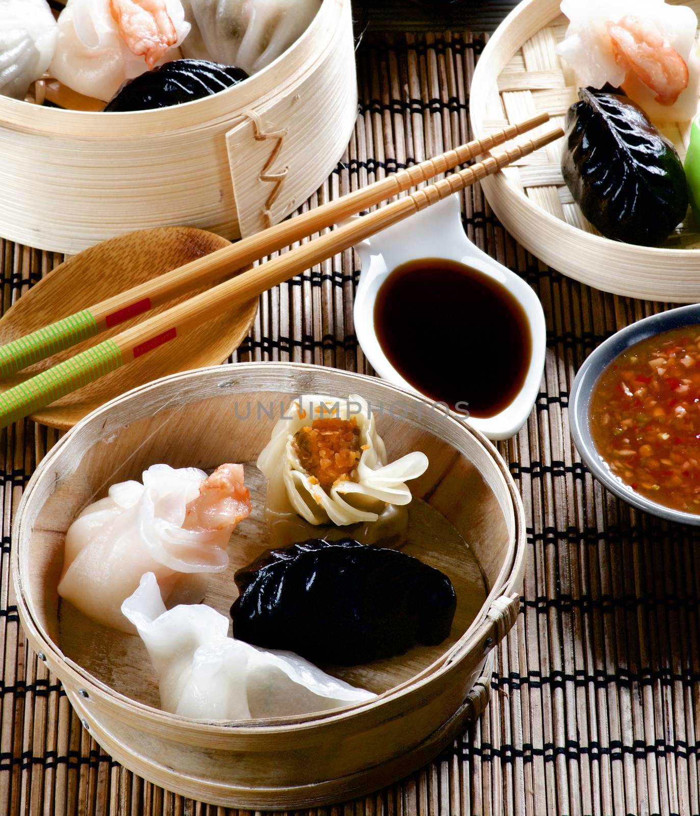 Assorted Dim Sum in Bamboo Steamed Bowls, Red Chili and Soy Sauces with Chopsticks closeup on Straw Mat background