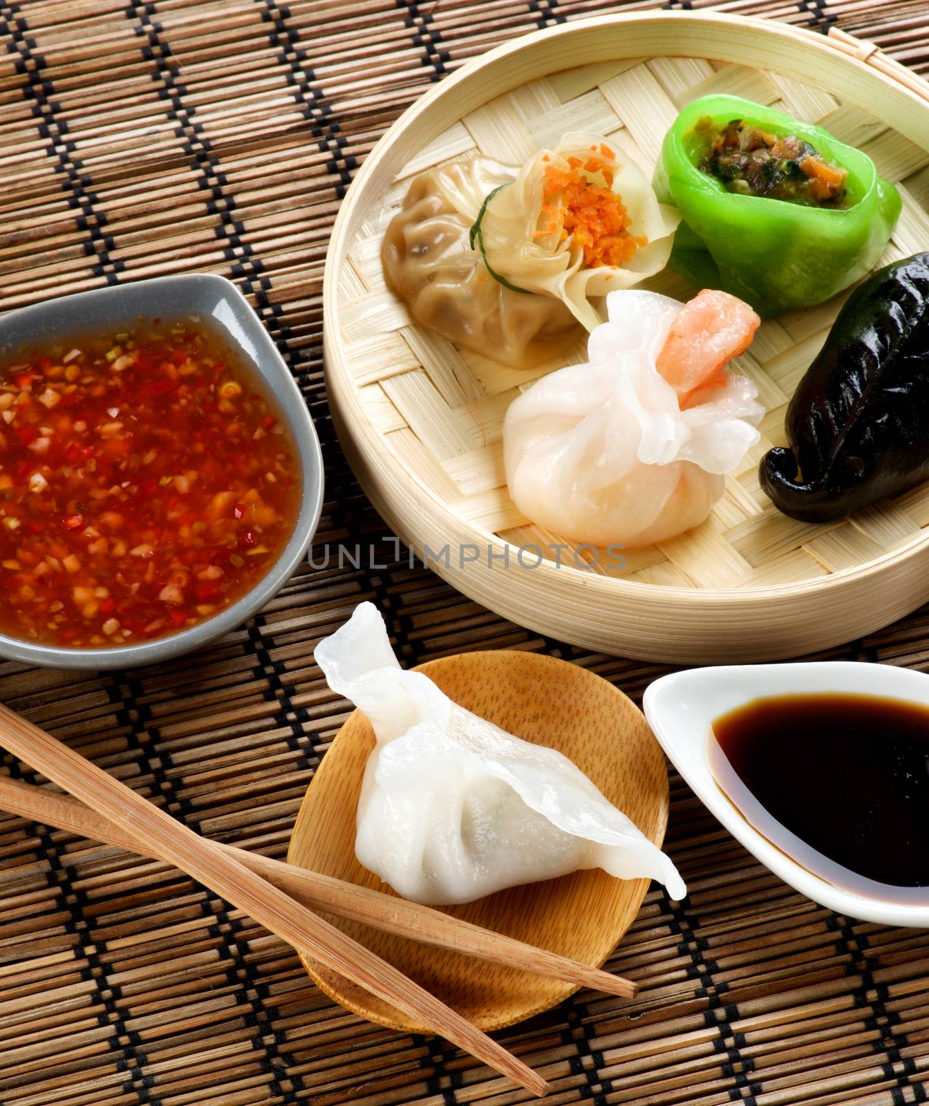 Assorted Dim Sum in Bamboo Steamed Bowl and Dai-Tory with Chiken on Wooden Plate with Red Chili and Soy Sauces and Chopsticks closeup on Straw Mat background