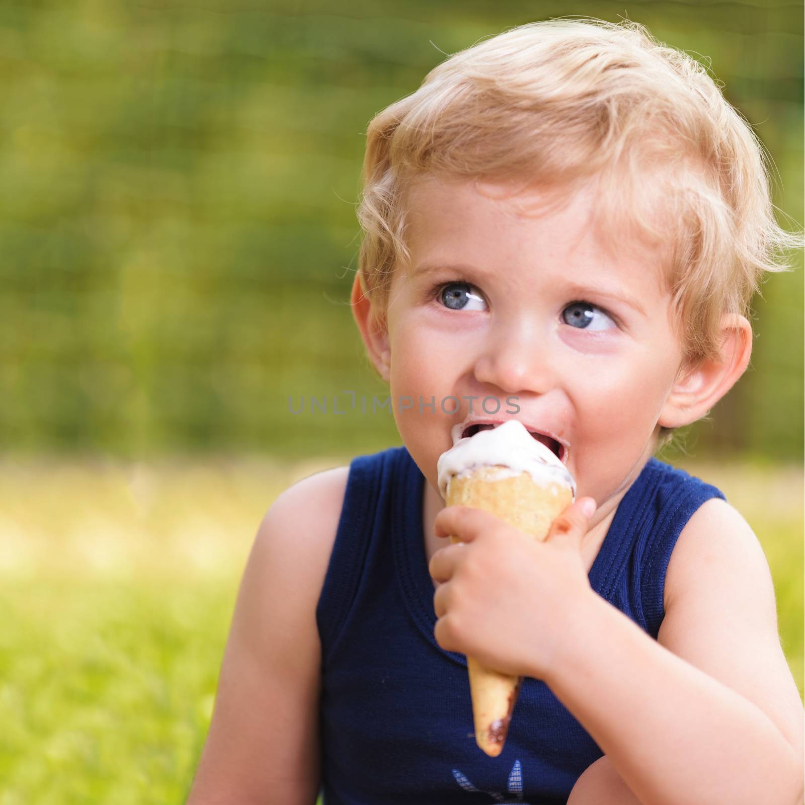 Sweet toddler baby boy eighteen months old, eating tasty ice cream over background with green bokeh outdoor.