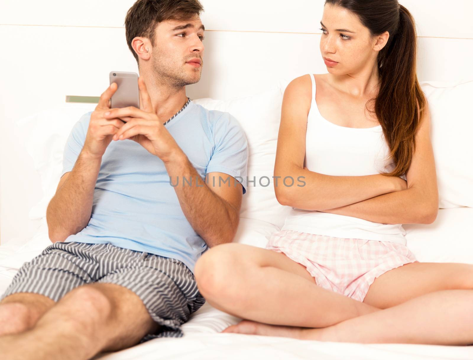 Addicted young man on bed with a cellphone while the woman looks angry