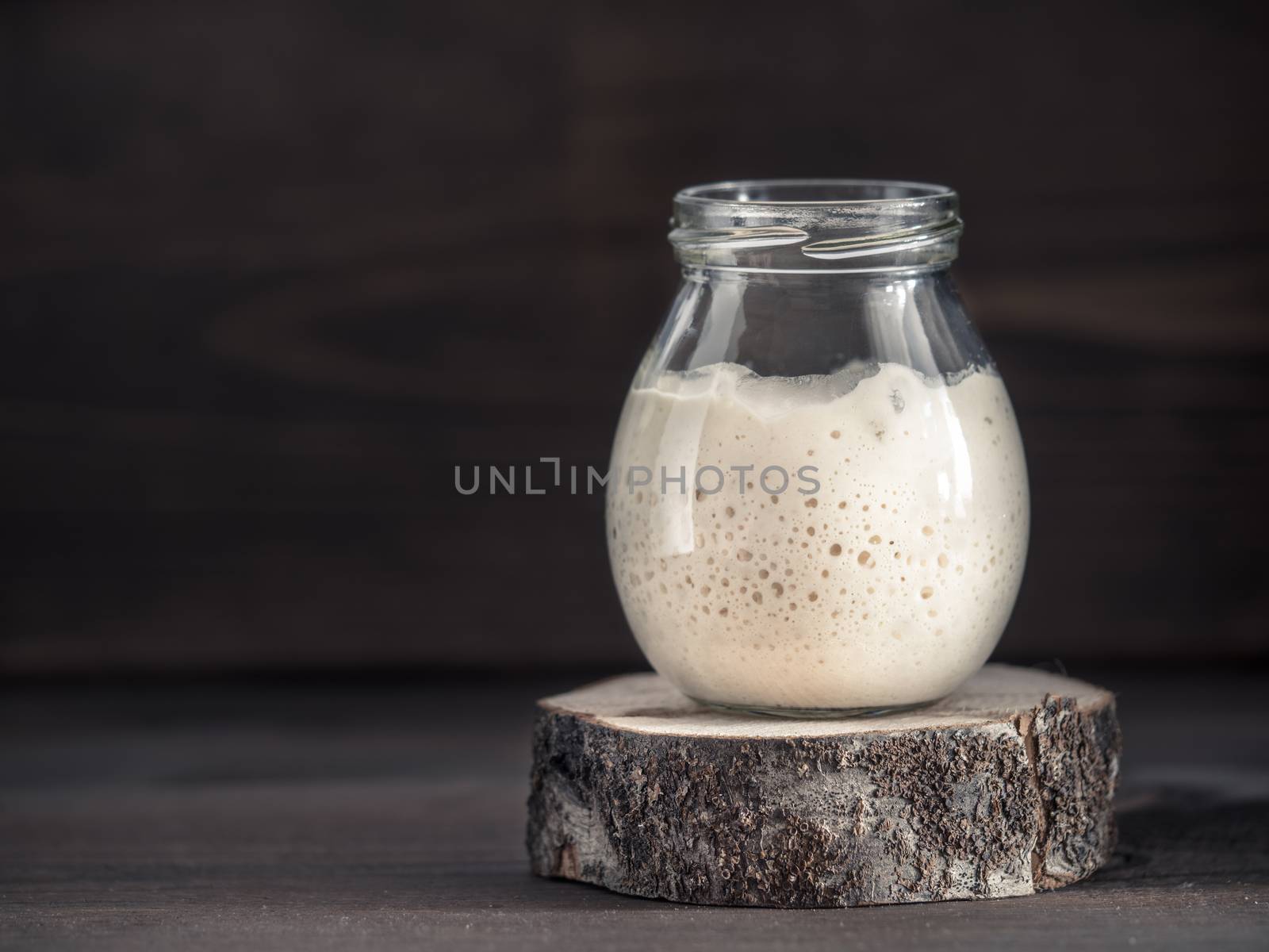 Active wheat sourdough starter in glass jar on brown wooden background. Starter for sourdough bread. Toned image. Copy space.