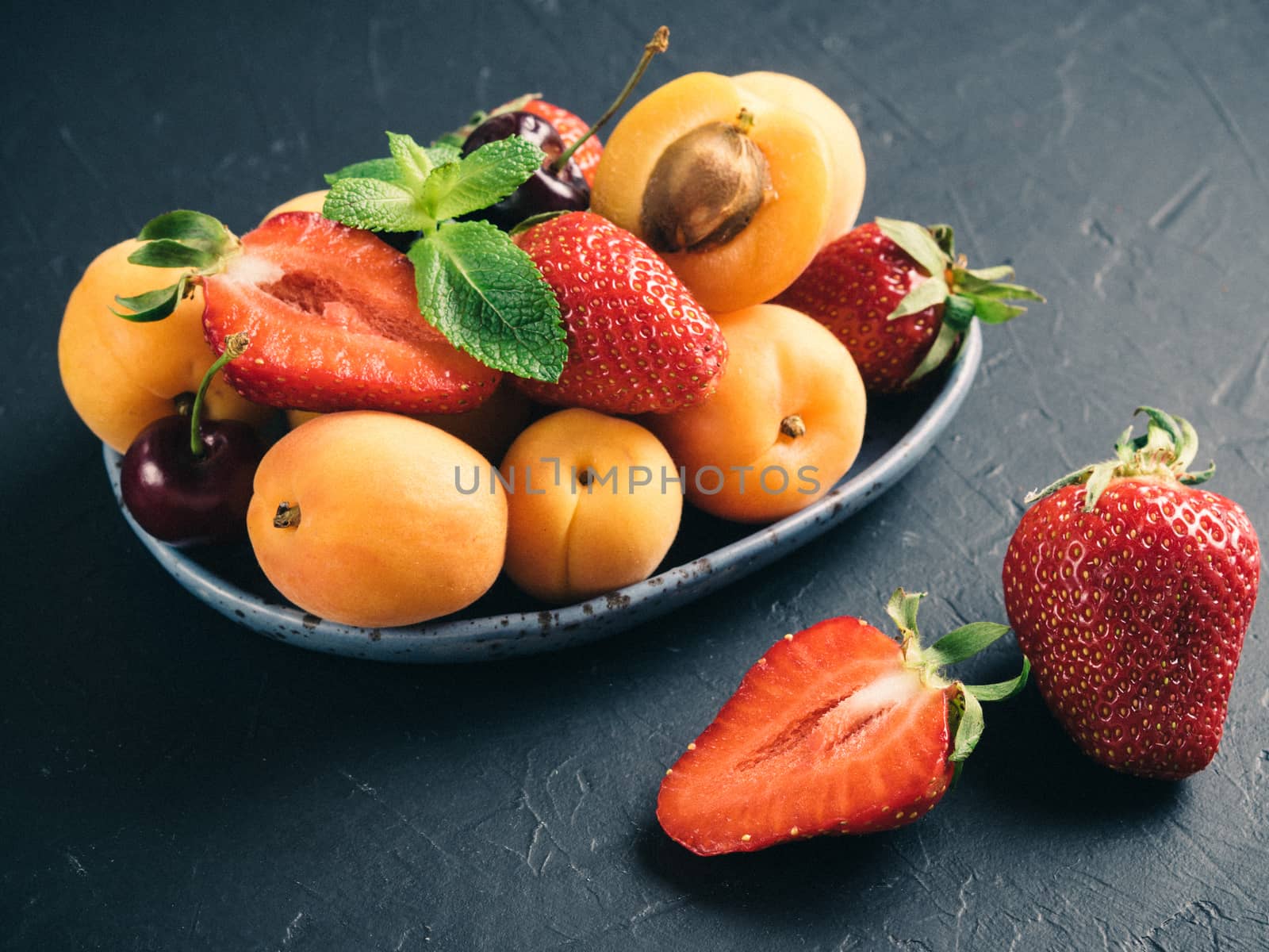 Closeup view of fruits and berries on dark background. Heap of fresh strawberries, blueberries, apricot and mint leaves in trendy plate. Focus on strawberry. Healthy food,superfood,diet,detox concept.