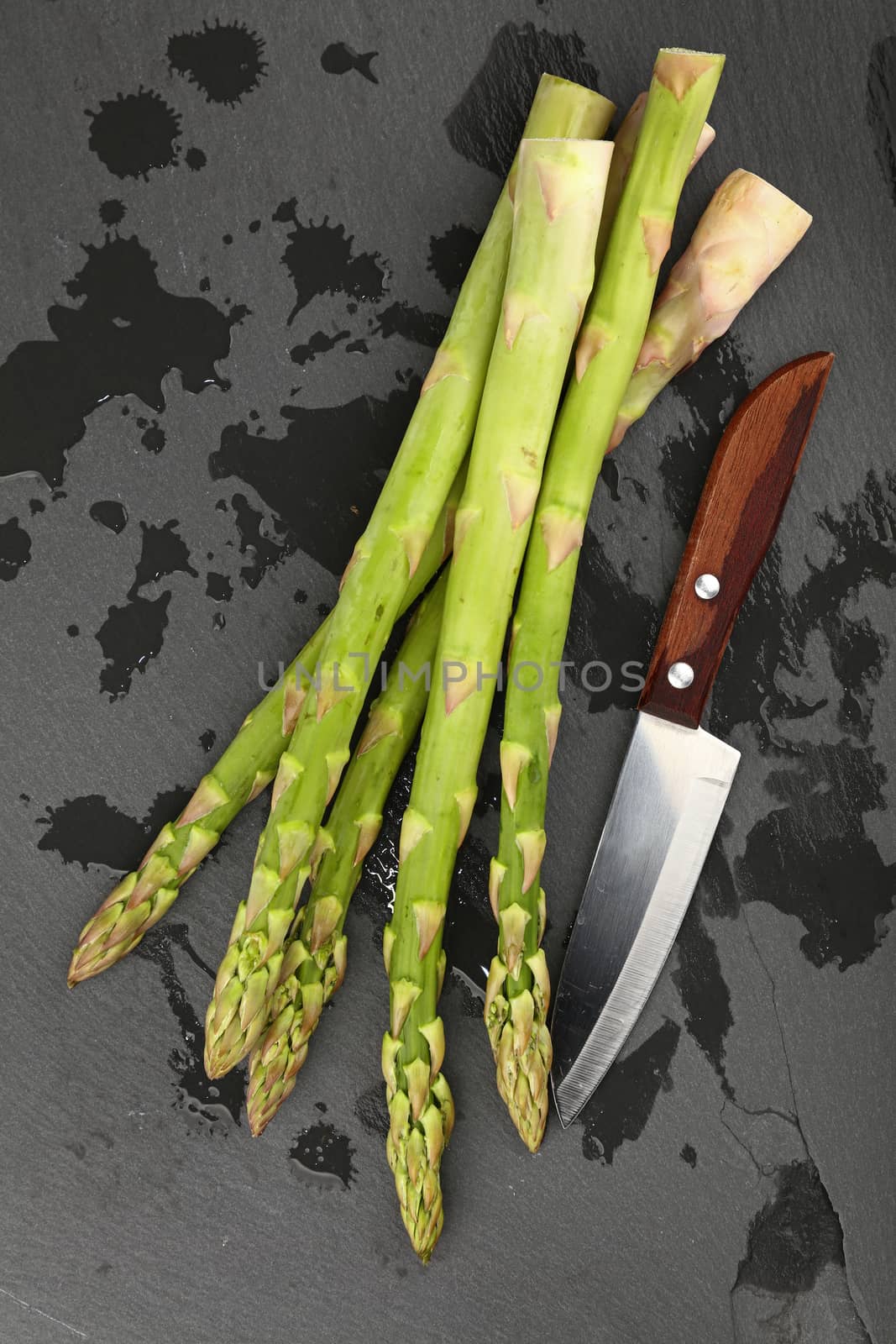 Fresh green washed asparagus and knife on black slate board with drops of water, close up, elevated top view