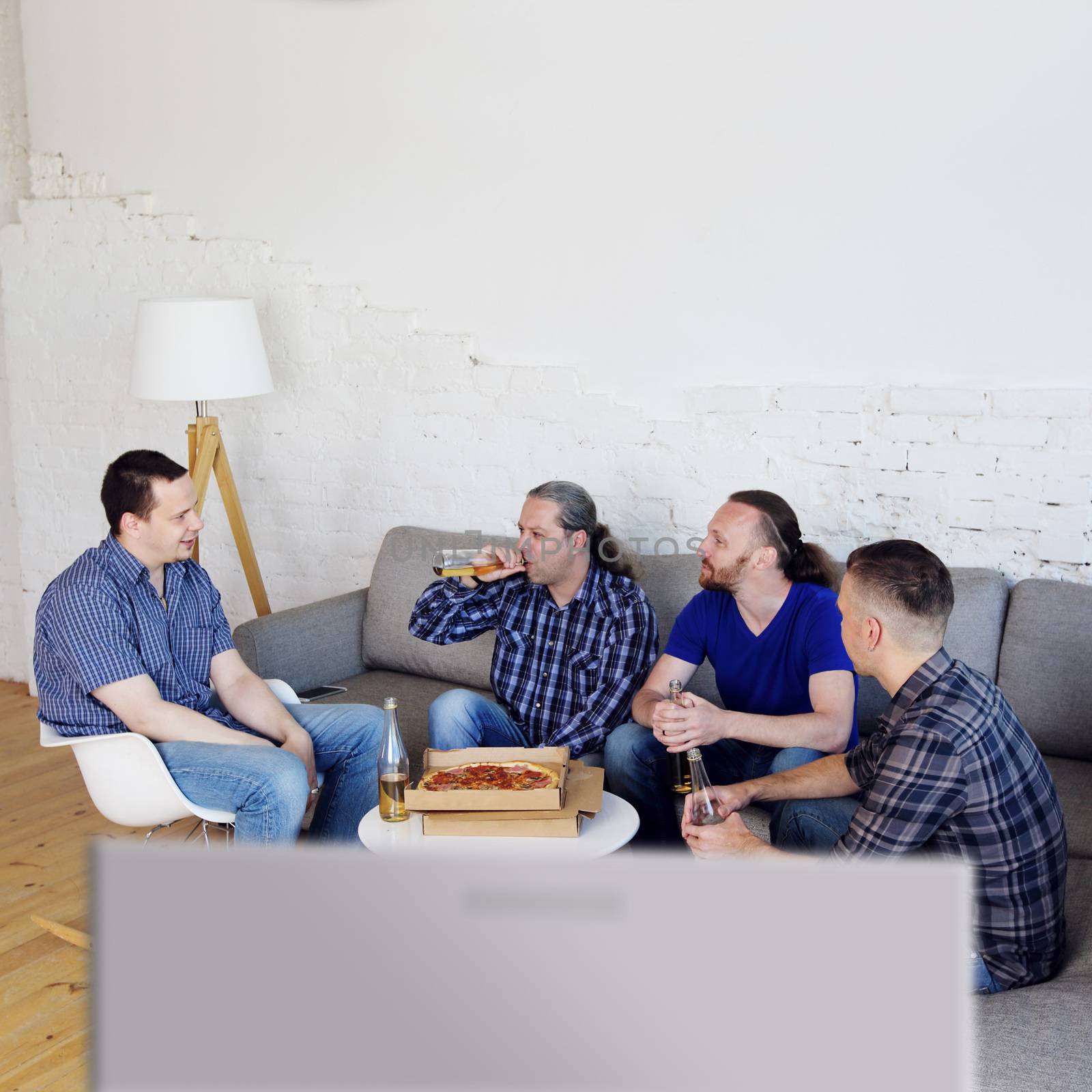 Group of men drinking beer, eating pizza, talking and smiling while resting at home on couch behind TV