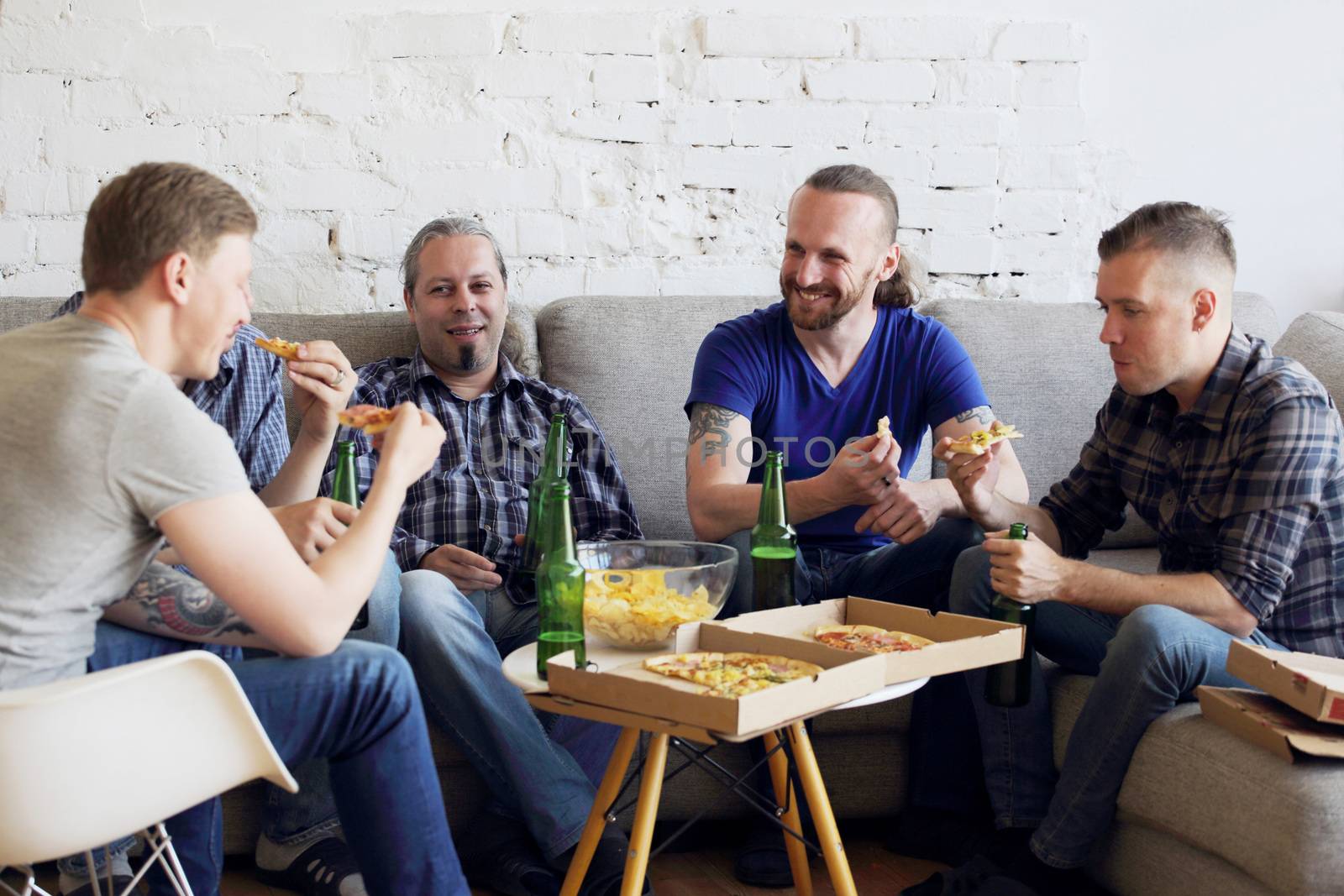 Group of men drinking beer, eating pizza, talking and smiling while resting at home on couch behind TV