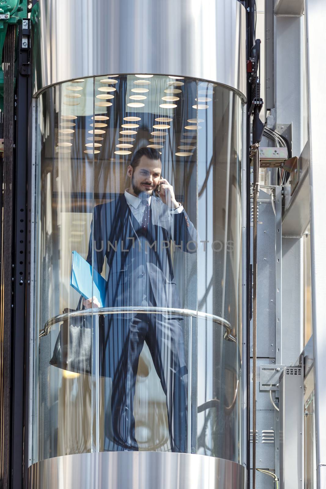 Businessman in modern glass elevator talking by the phone and holding papers