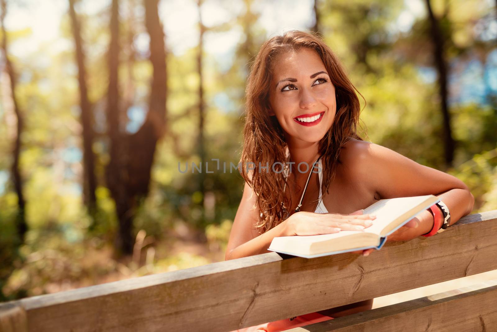 Young woman reading a book while relaxing in the sunny forest and looking away with smile on her thinking face.