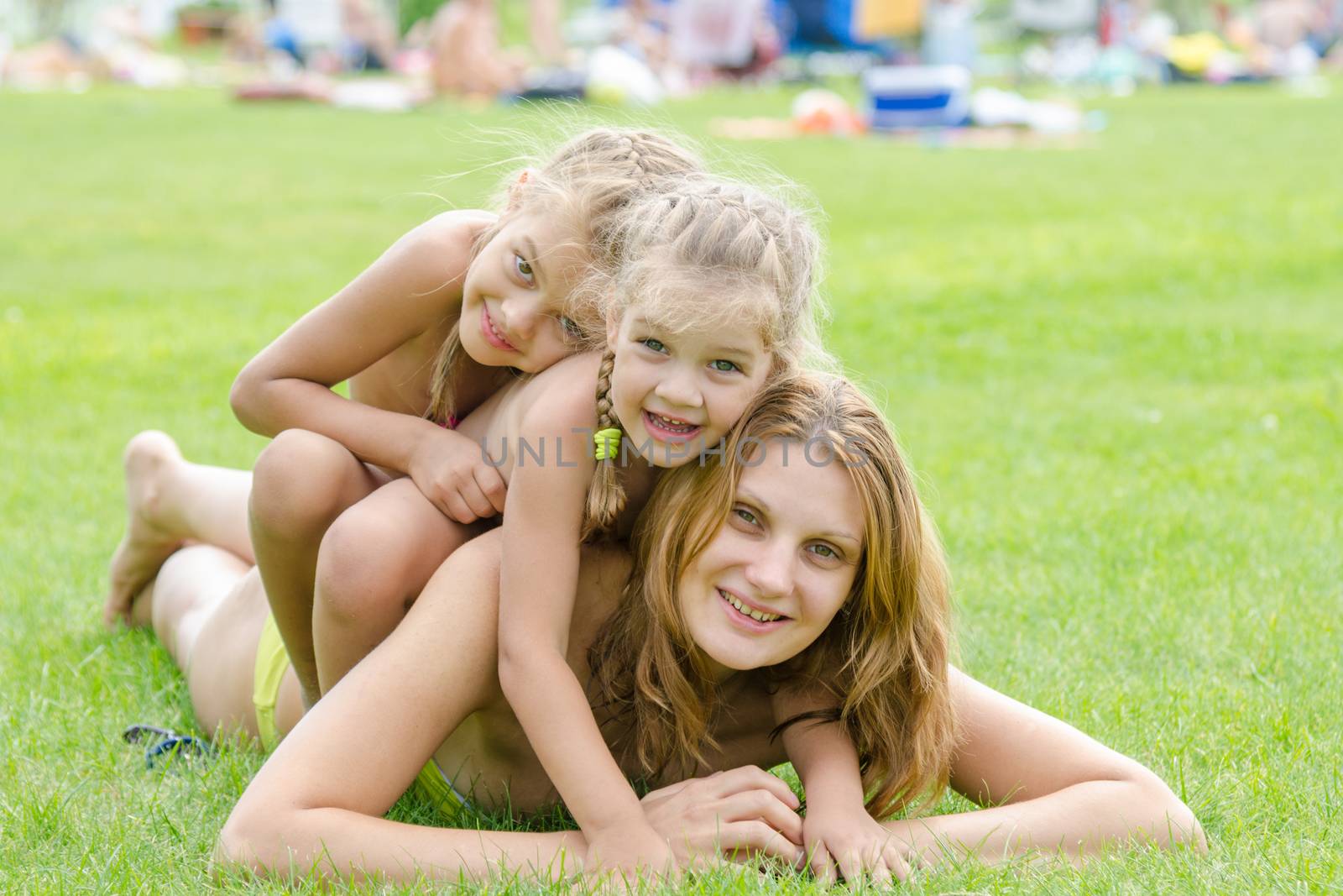 Two daughters happily sit on mom back, hot summer day relaxing on the green lawn