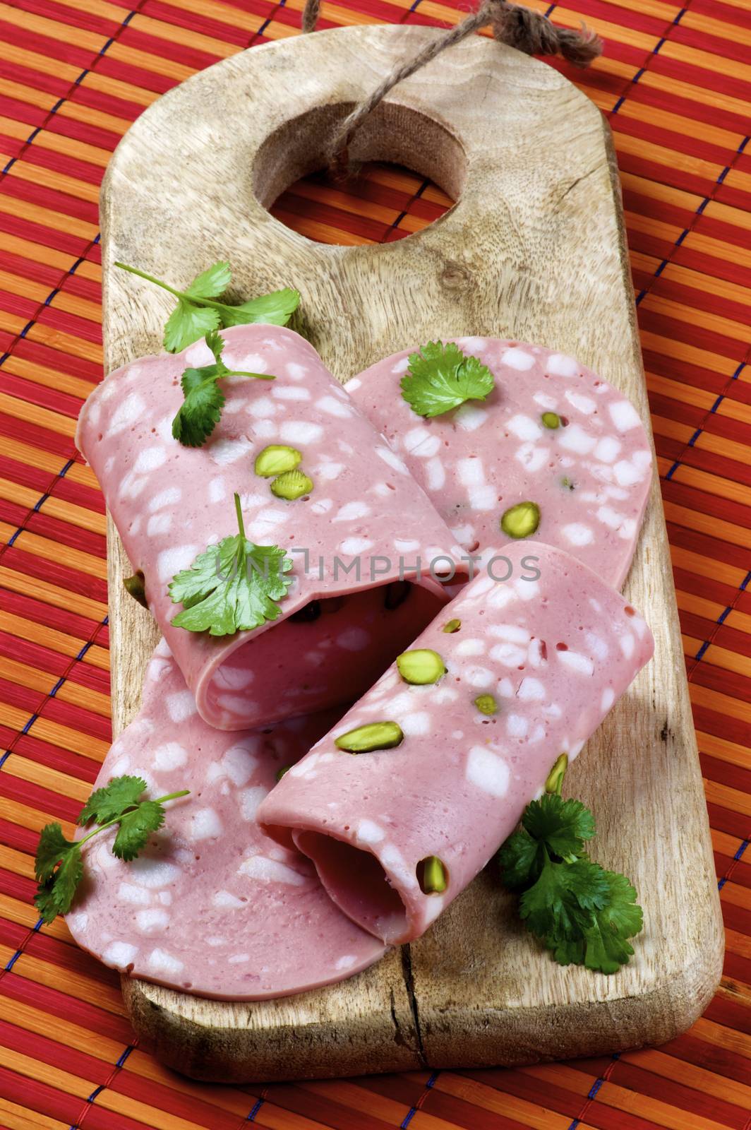 Slices of Delicious Fresh Mortadella with Pistachio and Greens on Wooden Cutting Board closeup on Straw mat background