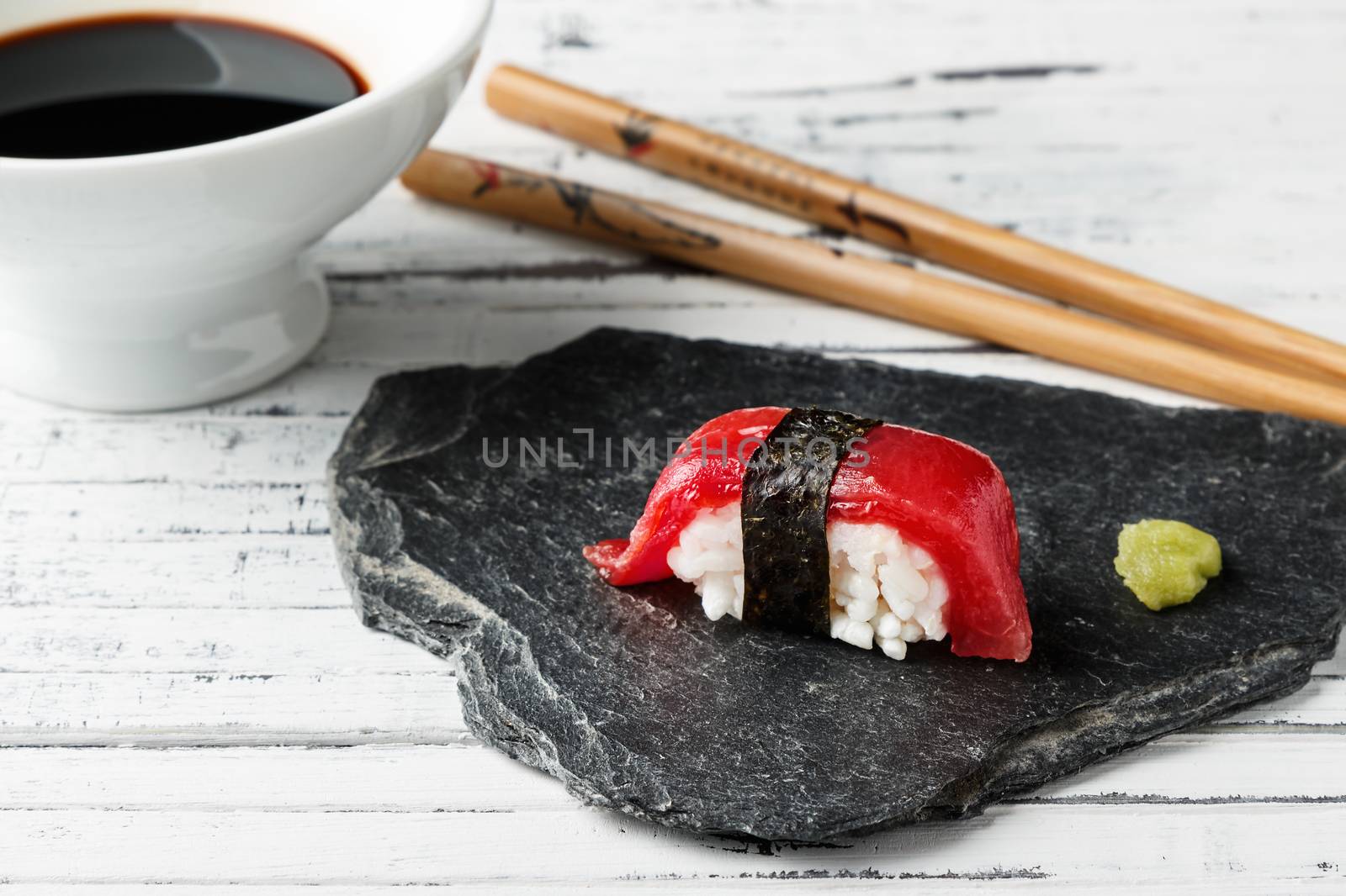 Red tuna Nigiri with Nori seaweed and wasabi paste on slate stone. Chopsticks and bowl with soy sauce in the background on old white wood. Raw fish in traditional Japanese sushi style. Horizontal image.