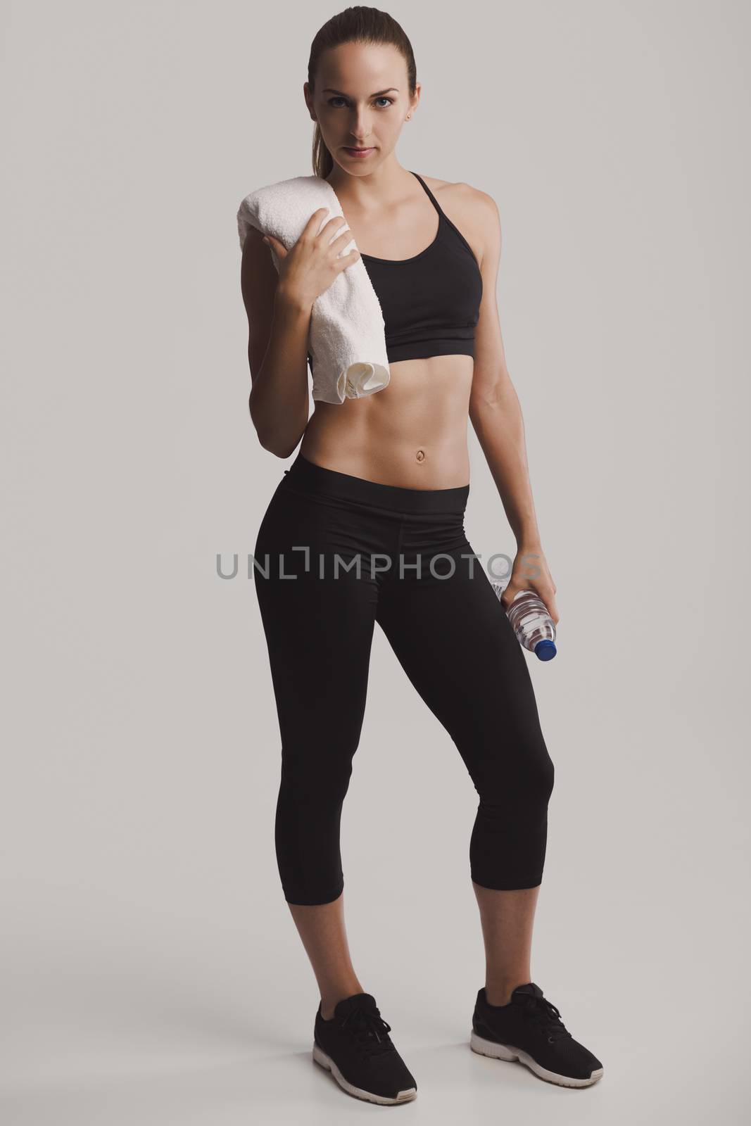 Portrait of sporty young woman holding a water bottle and towel, posing against a gray background