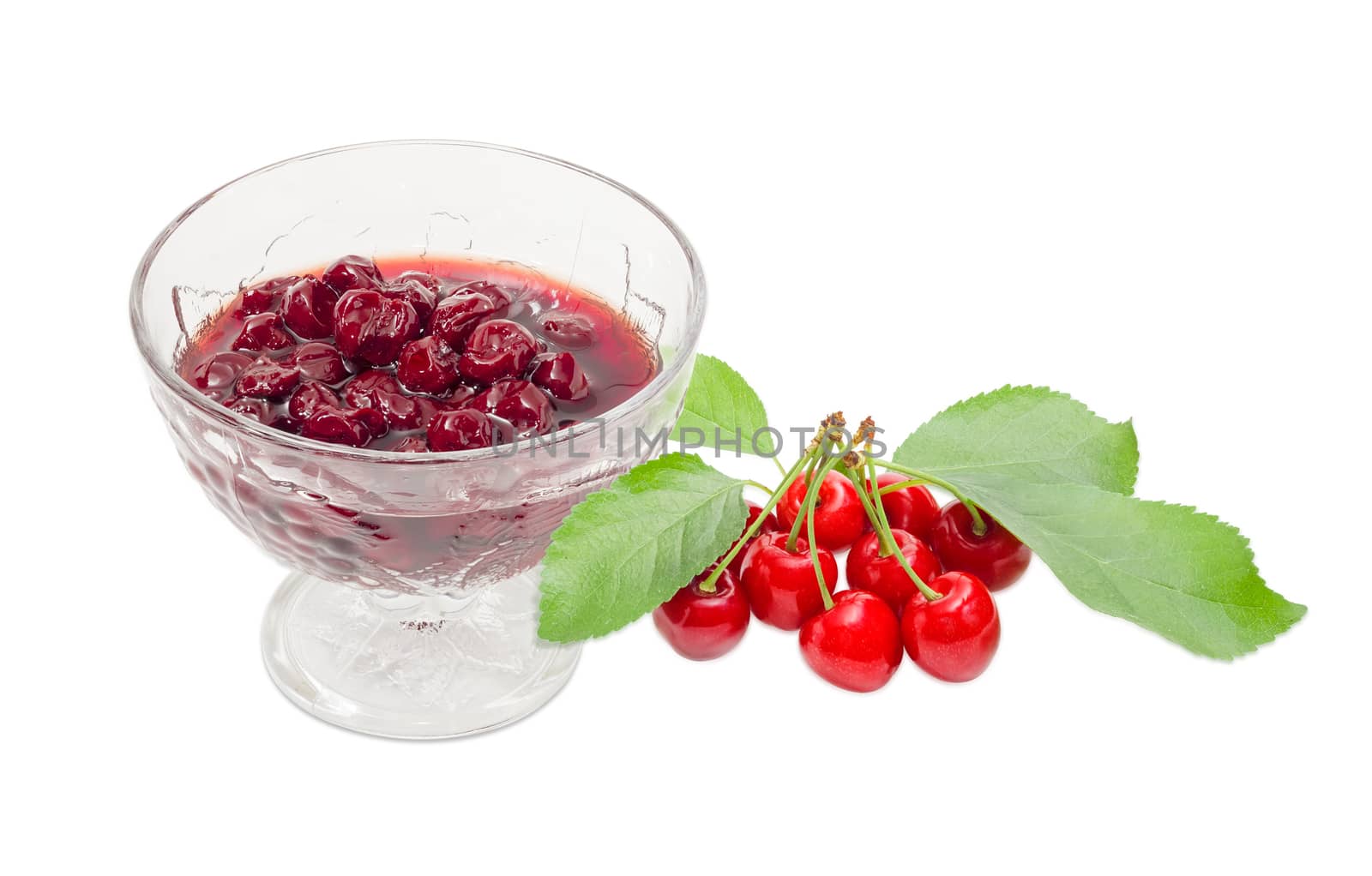 Cherry jam with whole fruits in the glass dessert stem bowl and several fresh cherries with the stalks and leaves beside on a light background
